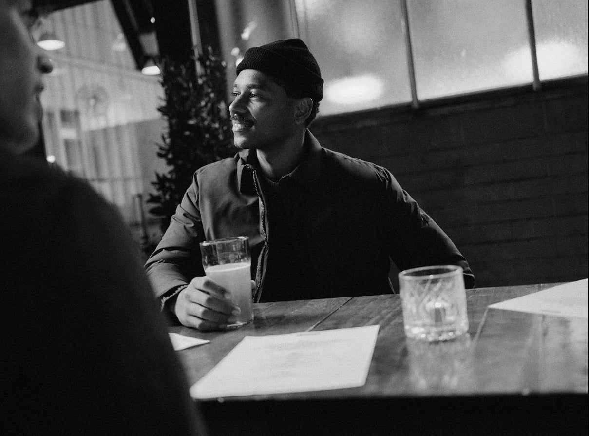 A man wearing a beanie and jacket sits at a wooden table outdoors at night, holding a glass of beer and looking to the side. Papers and another empty glass are on the table. The scene is in black and white.