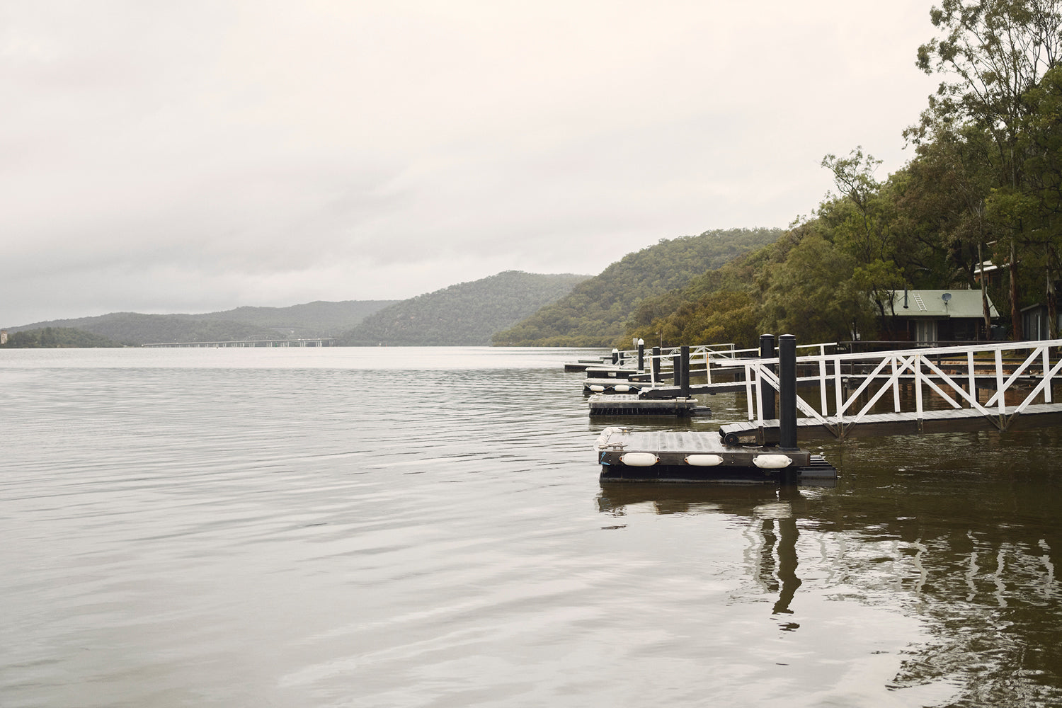 A calm lake with a floating dock and white railings, surrounded by green trees and hills under a cloudy, overcast sky. A small building is visible near the shoreline to the right.