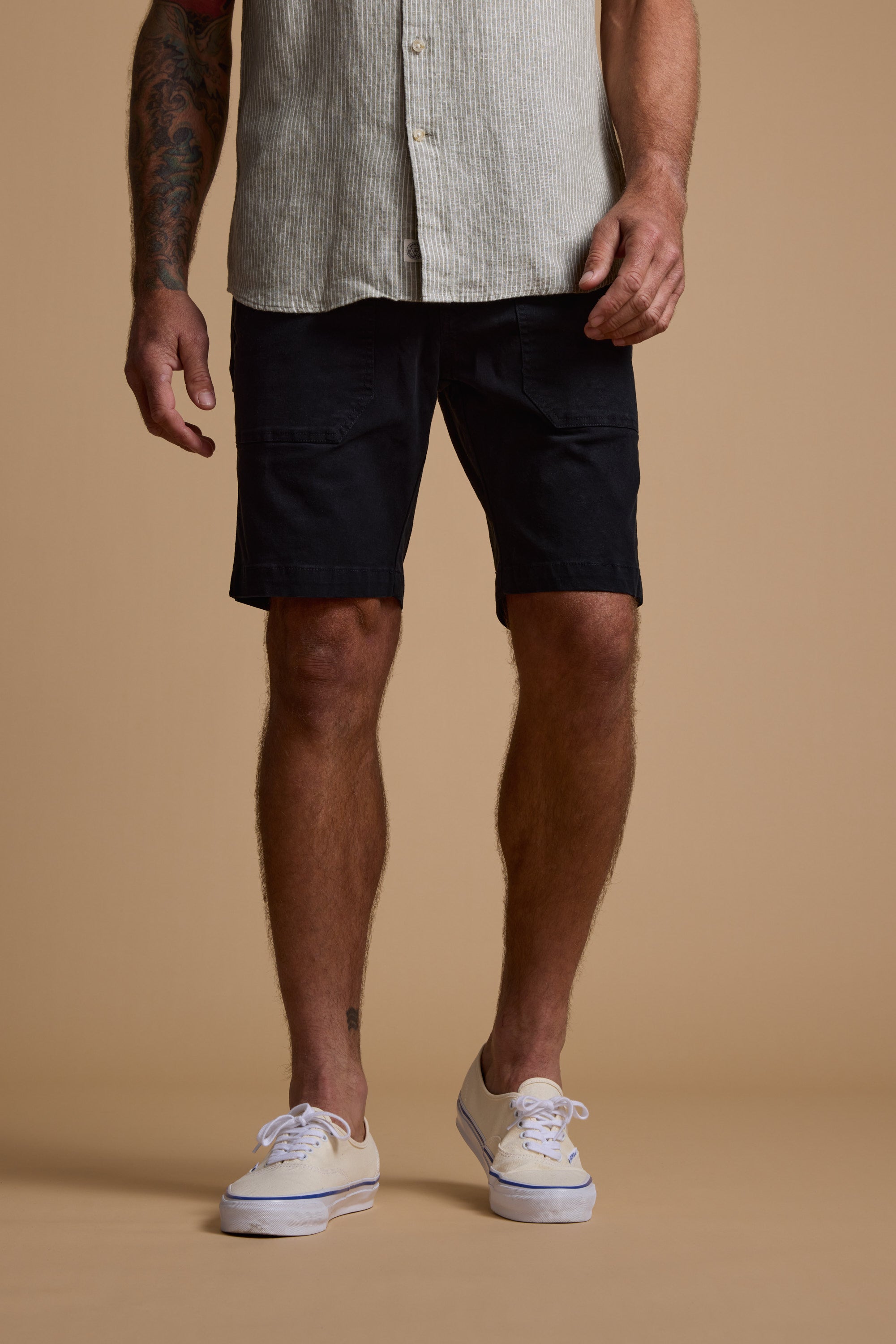 A man stands against a beige background wearing Barkers Tidal GD Utility Short in black, a light short-sleeve button-up shirt, and white sneakers. His arms are tattooed, and his face is not visible.