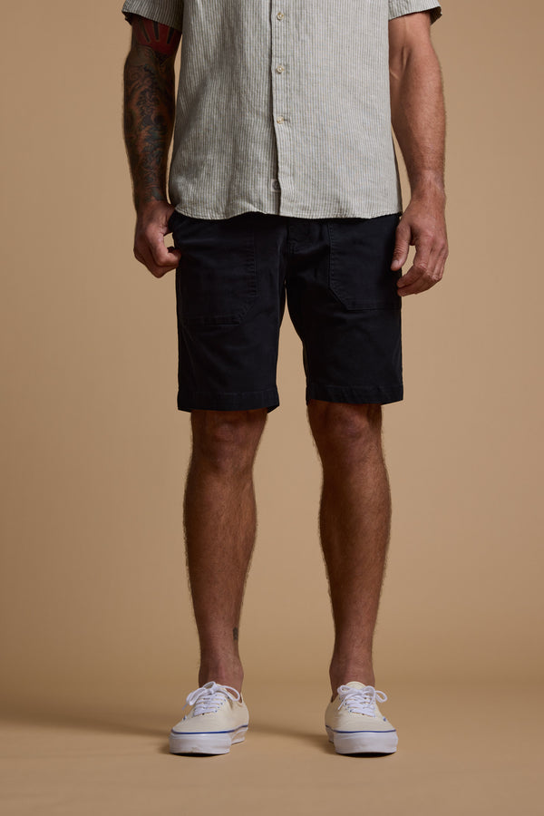A man stands against a plain beige background, shown from shoulders to feet, wearing a modern fit light striped button-up shirt, Barkers Tidal GD Utility Shorts in black, and white sneakers.