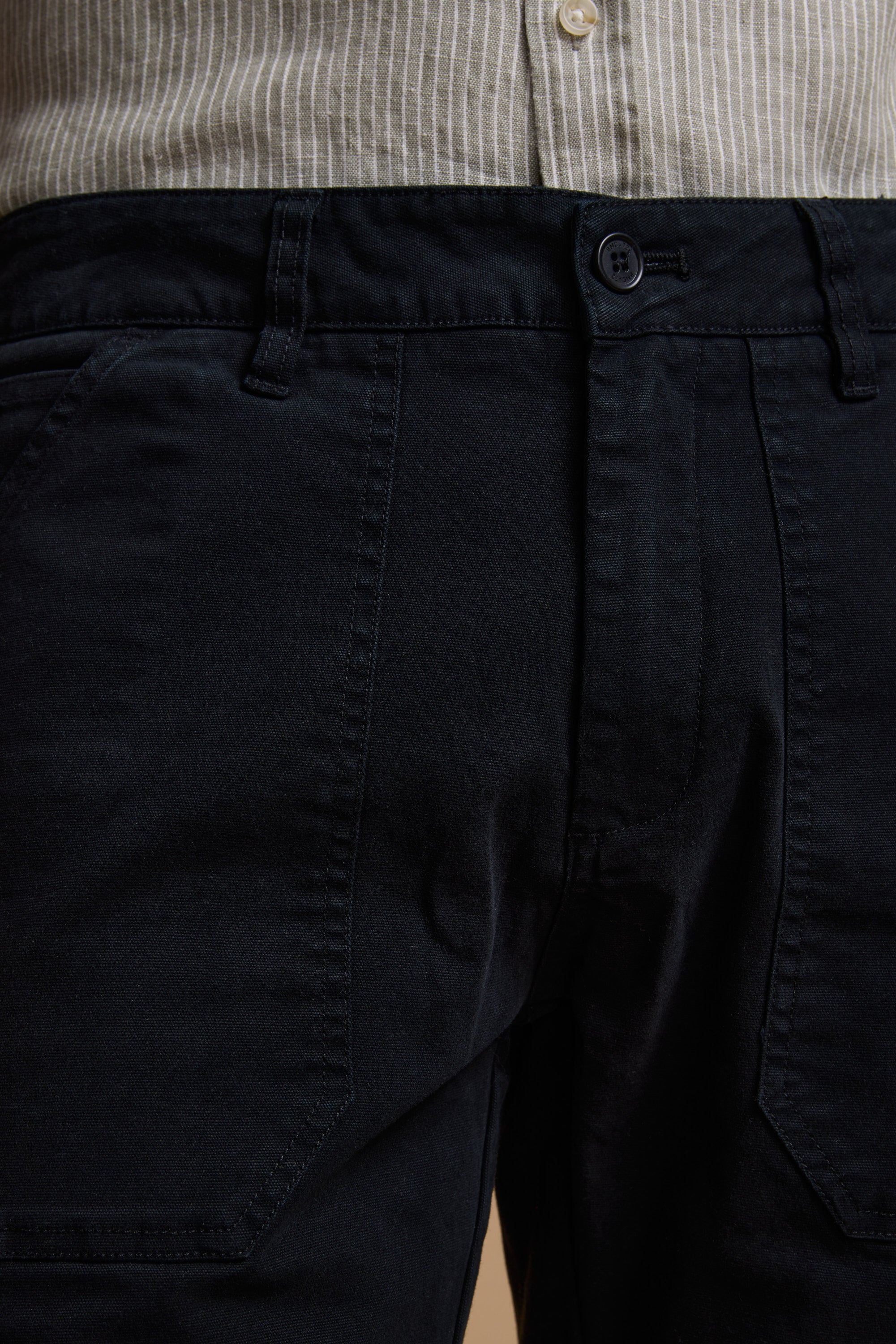 Close-up of a person wearing Barkers Tidal GD Utility Shorts in navy, featuring garment-dyed cotton, button and belt loops, paired with a beige and white striped shirt tucked in.