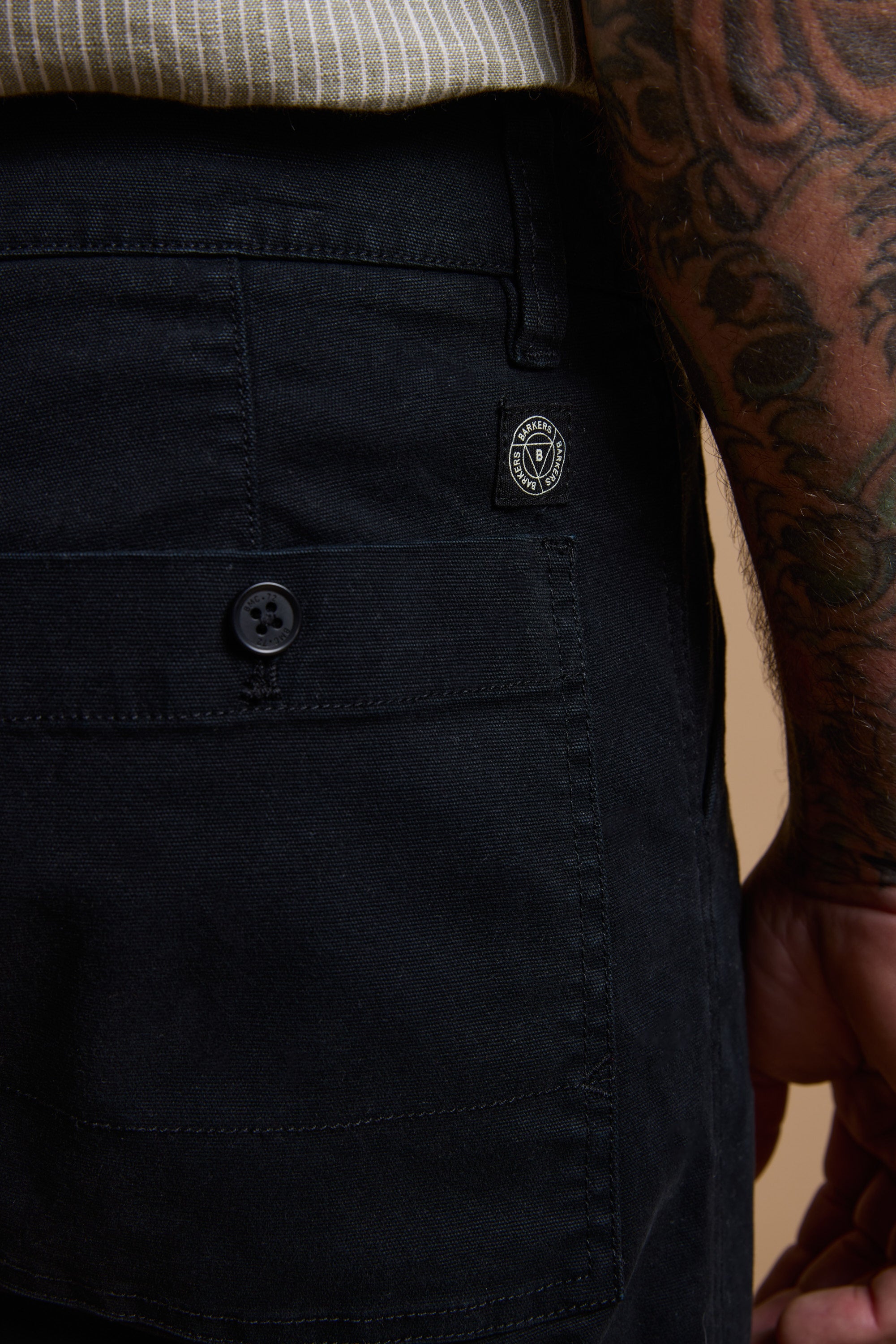 Close-up of a person wearing Barkers Tidal GD Utility Short—modern fit, black, with a buttoned pocket and small patch logo. The look is completed by a tattooed arm and striped shirt in comfortable garment-dyed cotton.