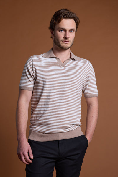 A man with light brown hair and a beard stands against a brown background, wearing the Barkers Parkway Cotton Stripe Polo in beige and white, paired with dark pants. He has one hand in his pocket and a neutral expression.