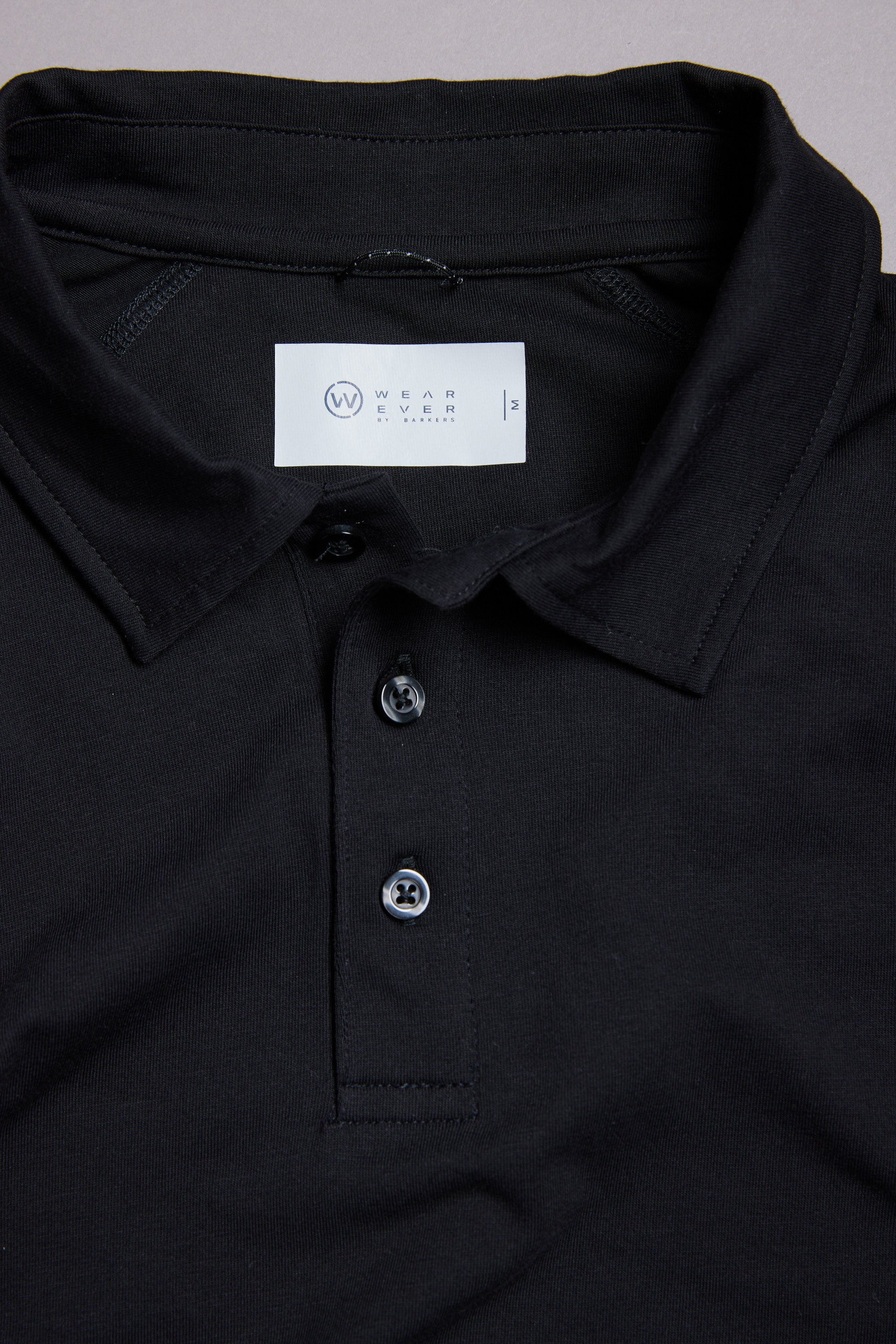 A close-up of the WearEver Outbound Polo in black, showcasing its collar, two buttons, and inner white label with WearEver branding and size info. Made from a breathable cotton-blend for lightweight stretch and all-day comfort.