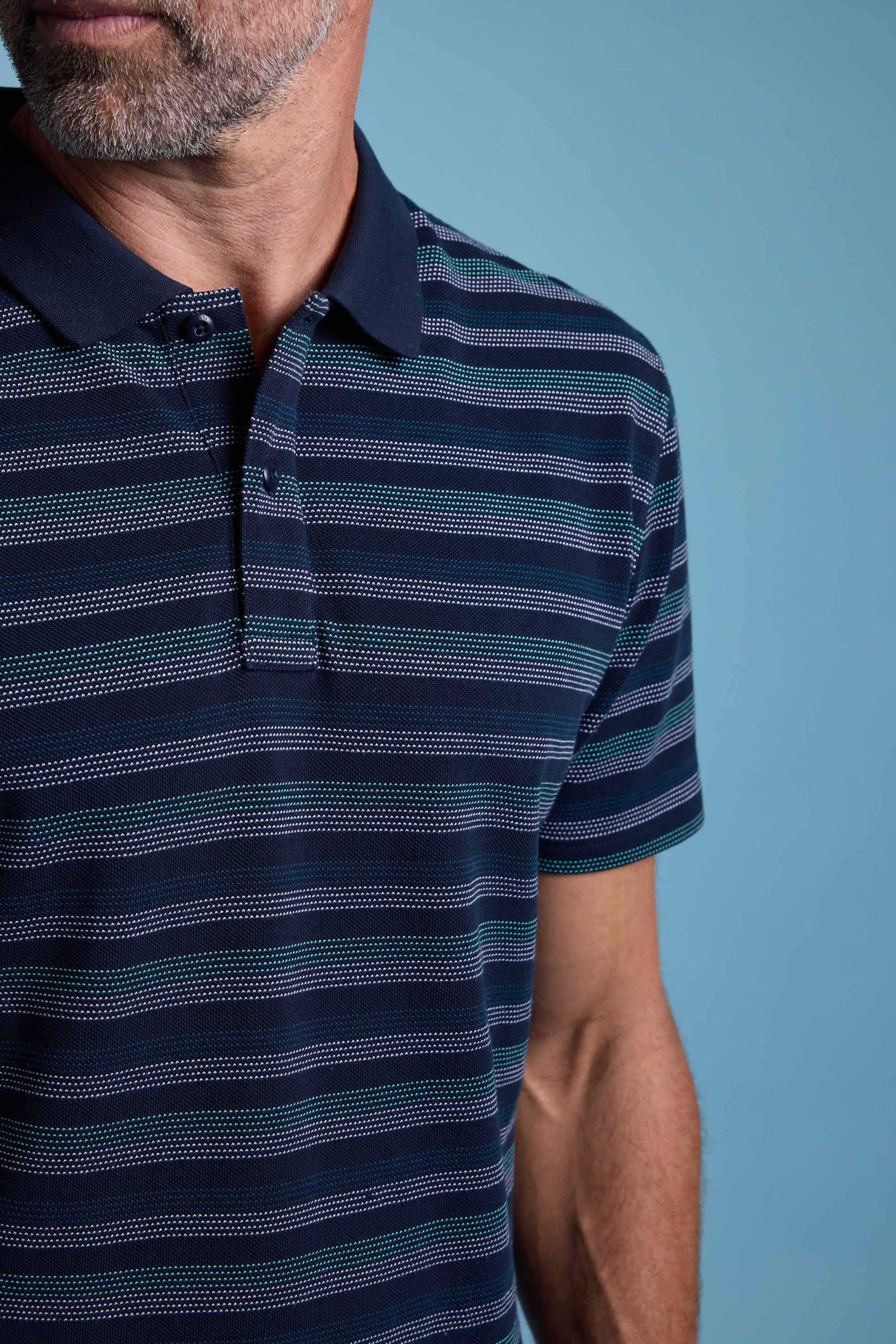 A close-up shows a man wearing the Barkers Dalton Stripe Polo in navy with teal and white stripes, made from organic cotton. Only his lower face and upper torso are visible against a light blue background—ideal for summer style.