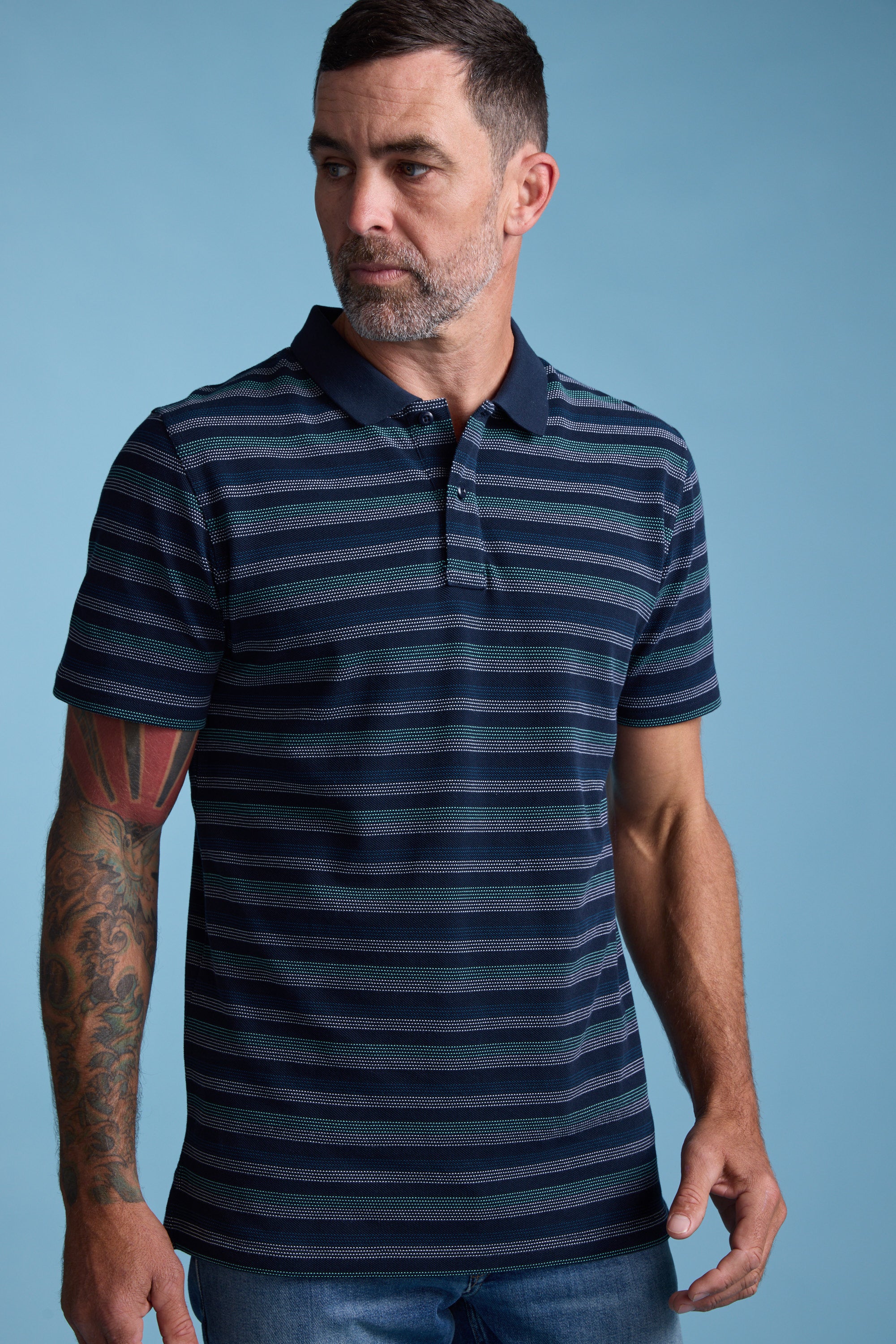A man with short dark hair and a beard wears the Barkers Dalton Stripe Polo with jeans, standing against a light blue background. His tattooed arm rests at his side as he looks right—a stylish pick for your summer wardrobe.