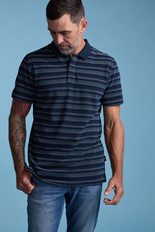 A man with short dark hair and a beard, wearing the Barkers Dalton Stripe Polo and blue jeans, stands against a light blue background, looking down with one hand on his shirt hem—a stylish summer essential.
