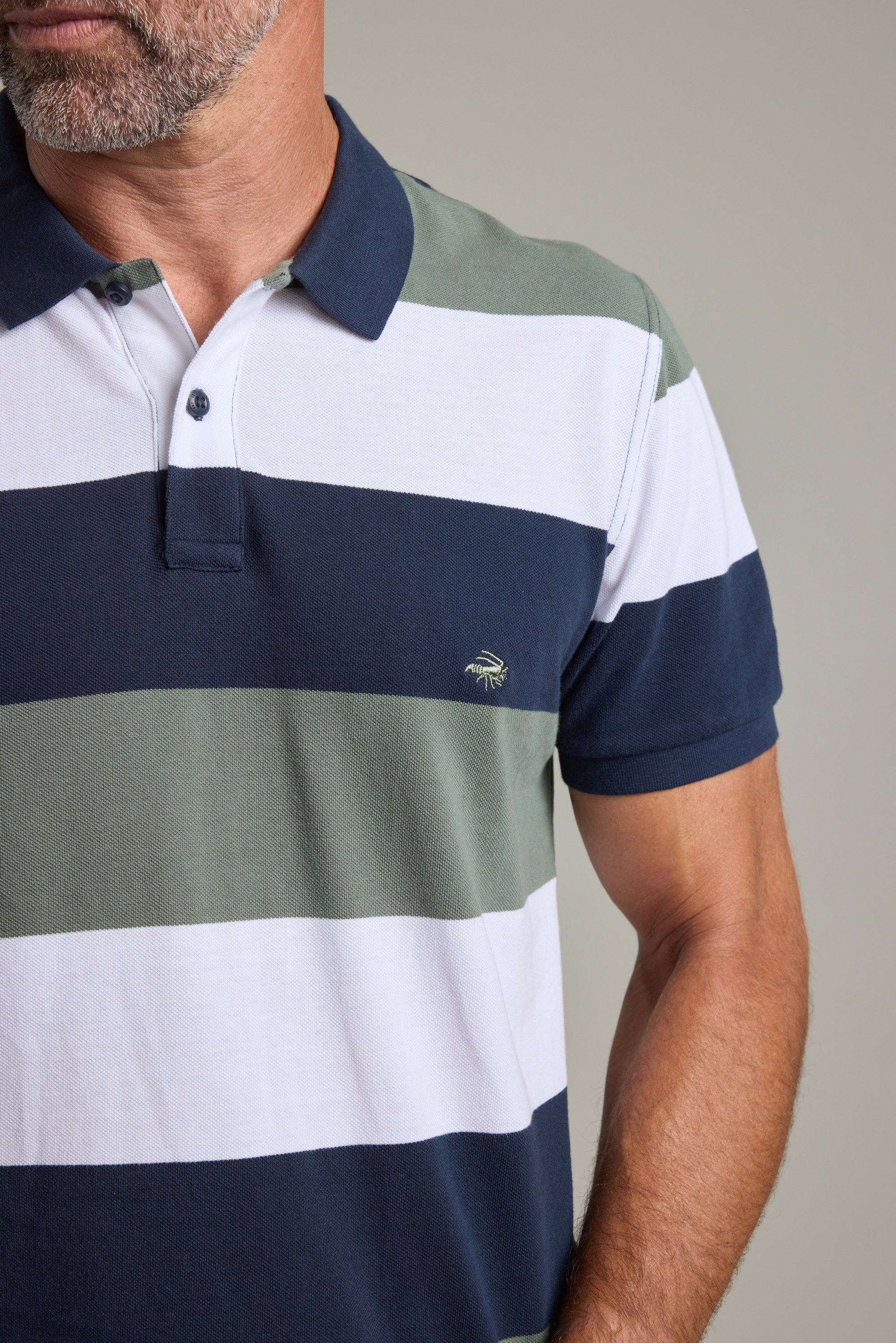 A man wears a Barkers Regent Pique Stripe Polo, a limited edition with short sleeves, wide white, green, and navy stripes, a collar, buttons, and an embroidered chest logo. The polo is partially out of frame.
