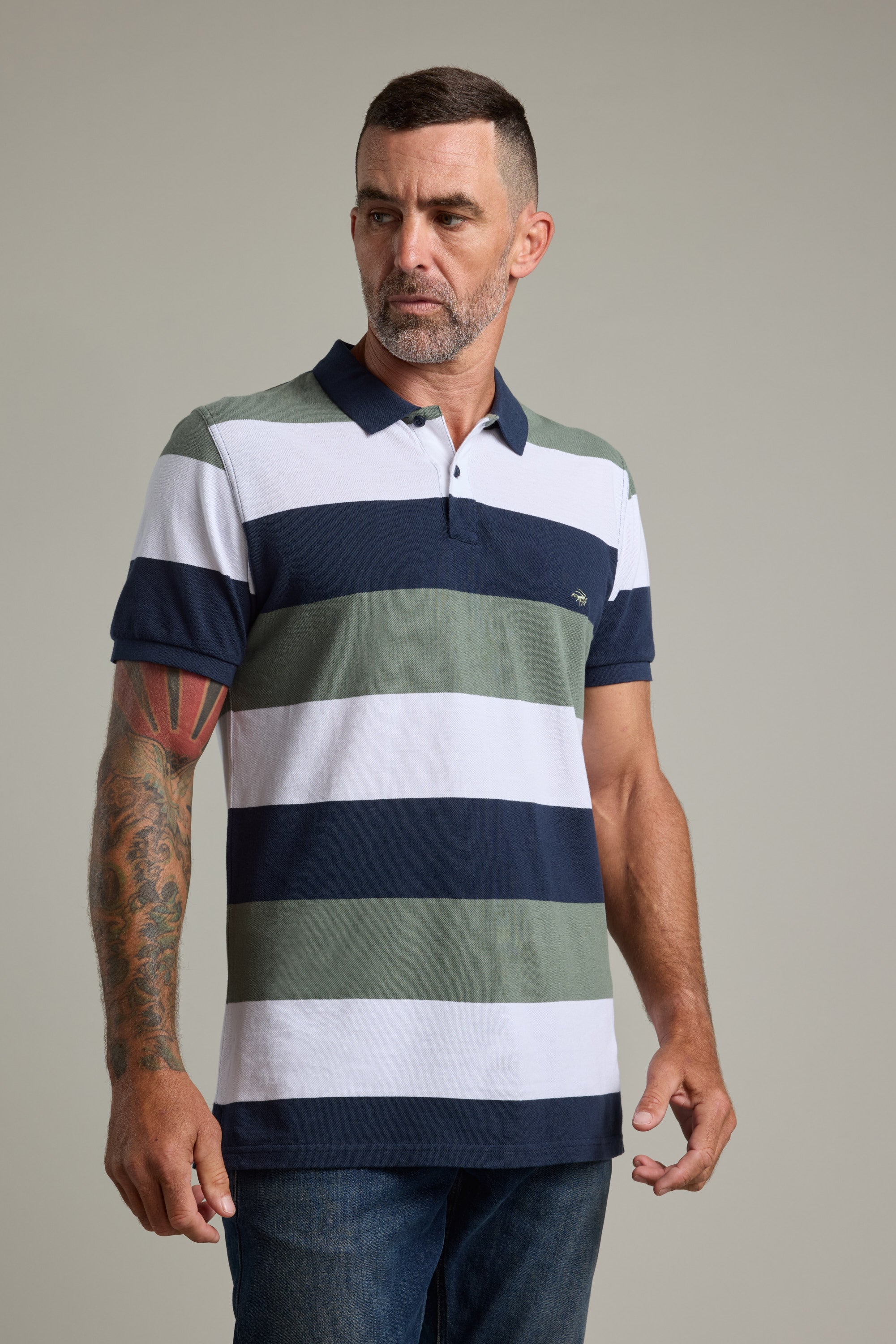 A man with short dark hair, a beard, and a colorful arm tattoo wears the Barkers Regent Pique Stripe Polo, featuring wide navy, green, and white stripes. He stands against a plain light background, looking to the side.