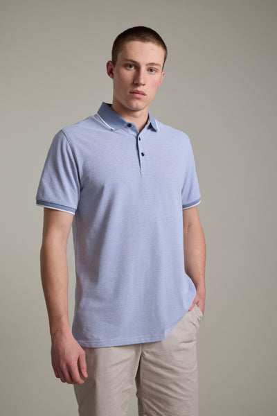 A young man with short hair wears the Barkers Birdie Pique Polo in light blue and beige shorts, standing against a plain, light-colored background with one hand in his pocket.