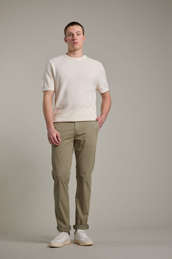 A man stands against a gray background, wearing an off-white short-sleeve sweater, Barkers Kingly Chino Pants made from organic cotton, and white sneakers. His expression is neutral with hands by his sides.
