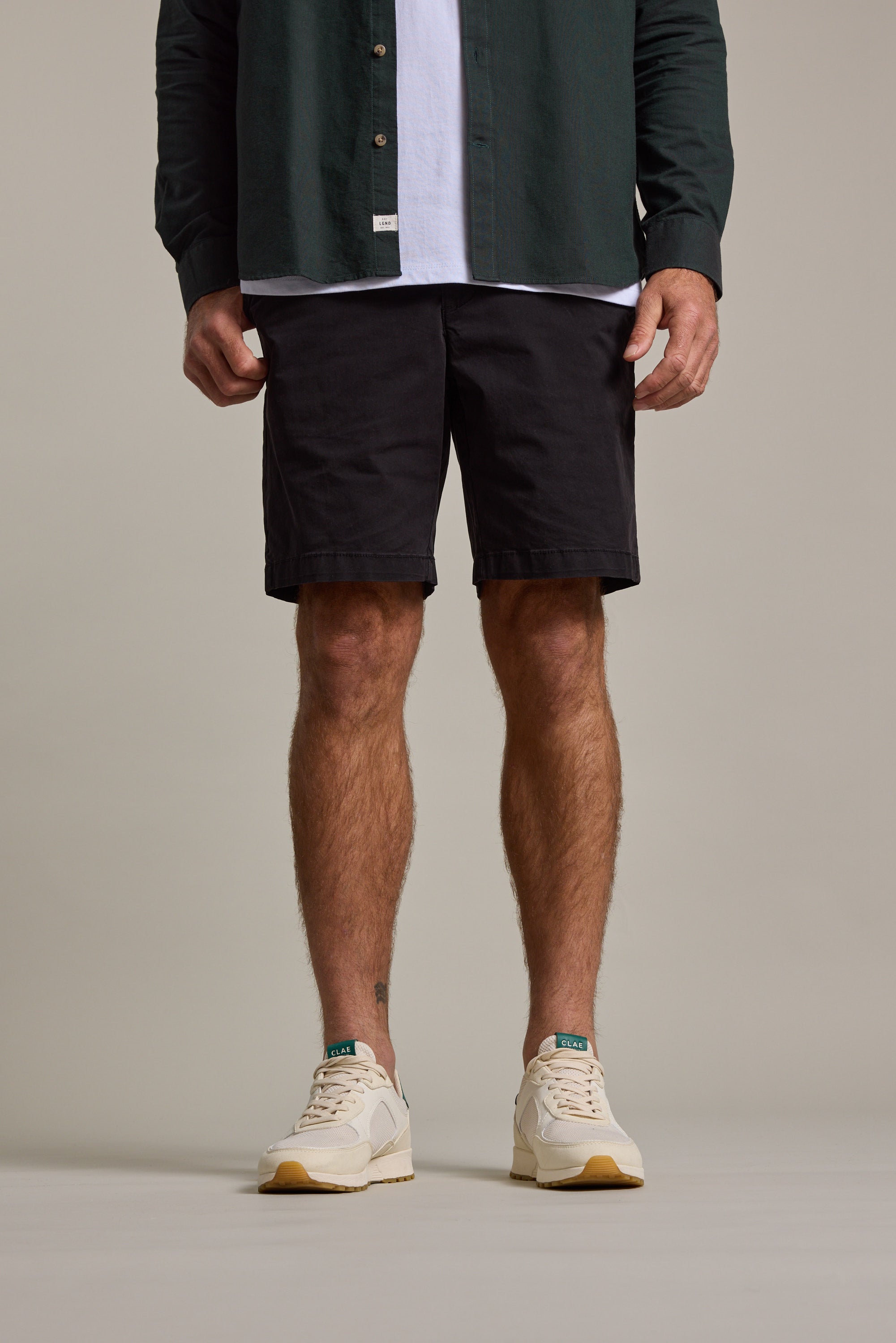 A person wears Barkers Kingly Chino Short men's stretch shorts paired with beige sneakers and a dark green shirt over a white t-shirt, standing against a plain background. The cropped image shows their body from the waist down.