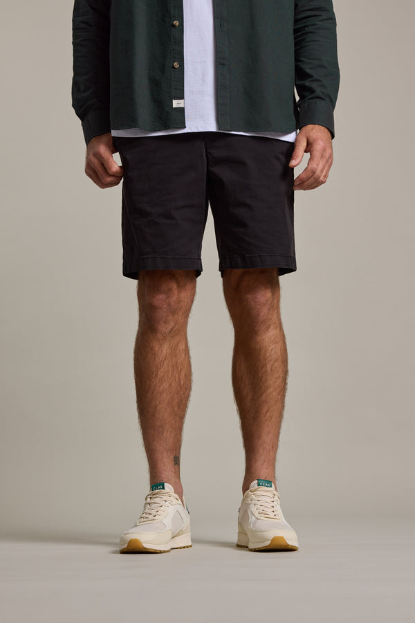 A person wears Barkers Kingly Chino Short men's stretch shorts paired with beige sneakers and a dark green shirt over a white t-shirt, standing against a plain background. The cropped image shows their body from the waist down.