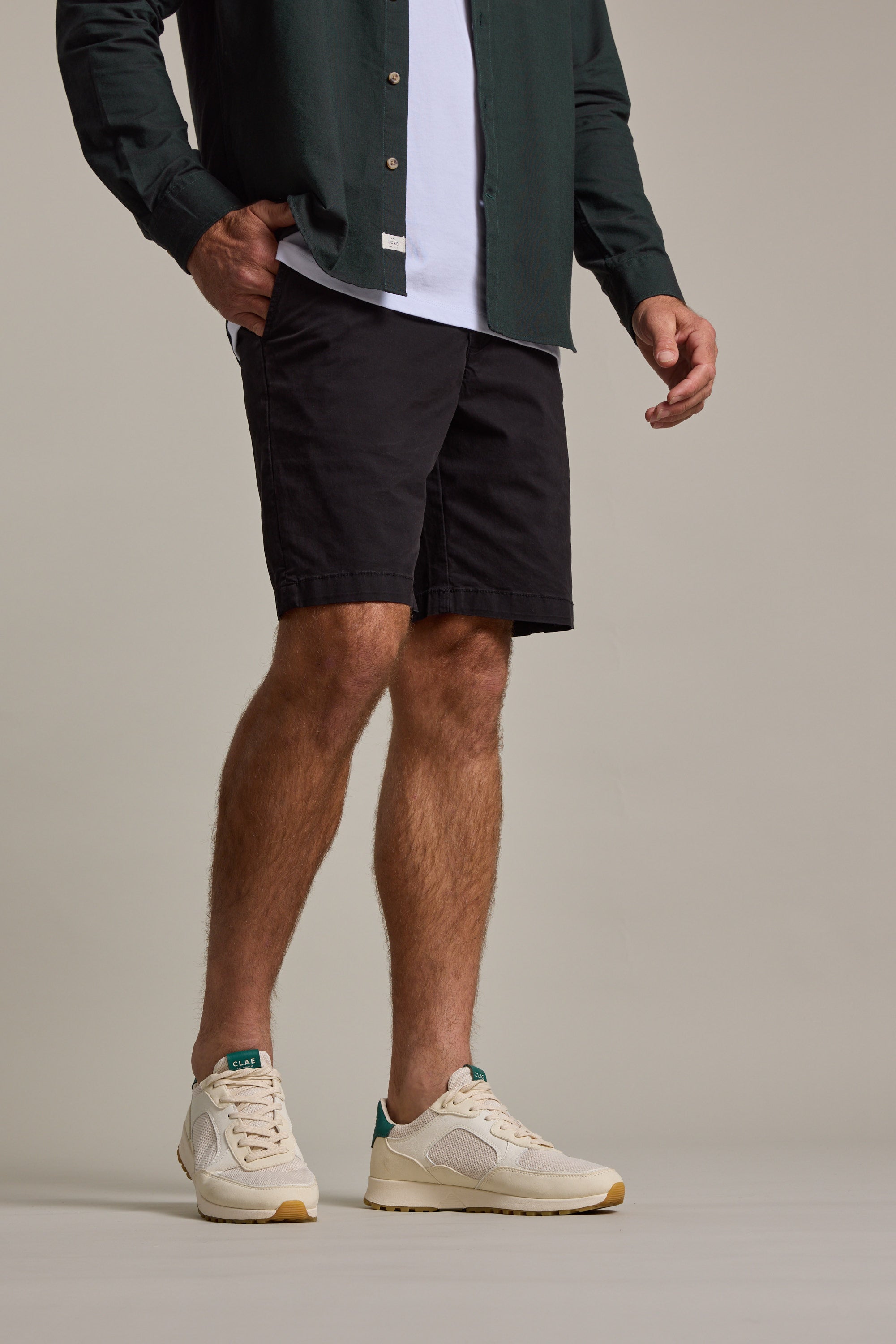 A man stands with one hand in his pocket, wearing a dark green shirt, white tee, and Barkers Kingly Chino Short—men's stretch shorts made from organic cotton—with cream sneakers featuring green accents, set against a plain light background.