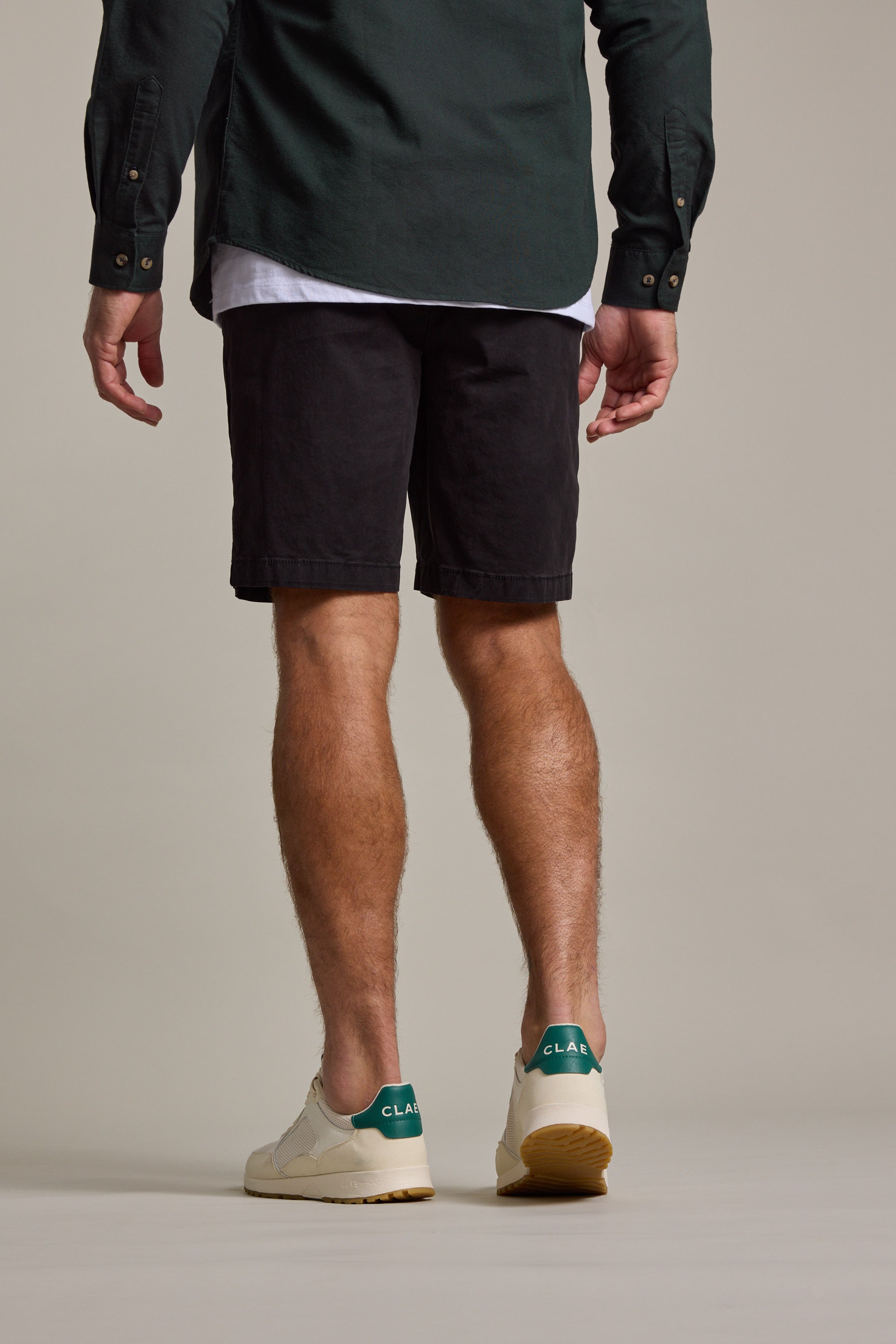 A man stands facing away, wearing a dark green button-up, white tee, Barkers Kingly Chino Shorts in black, and white sneakers with green heel accents against a plain beige background.