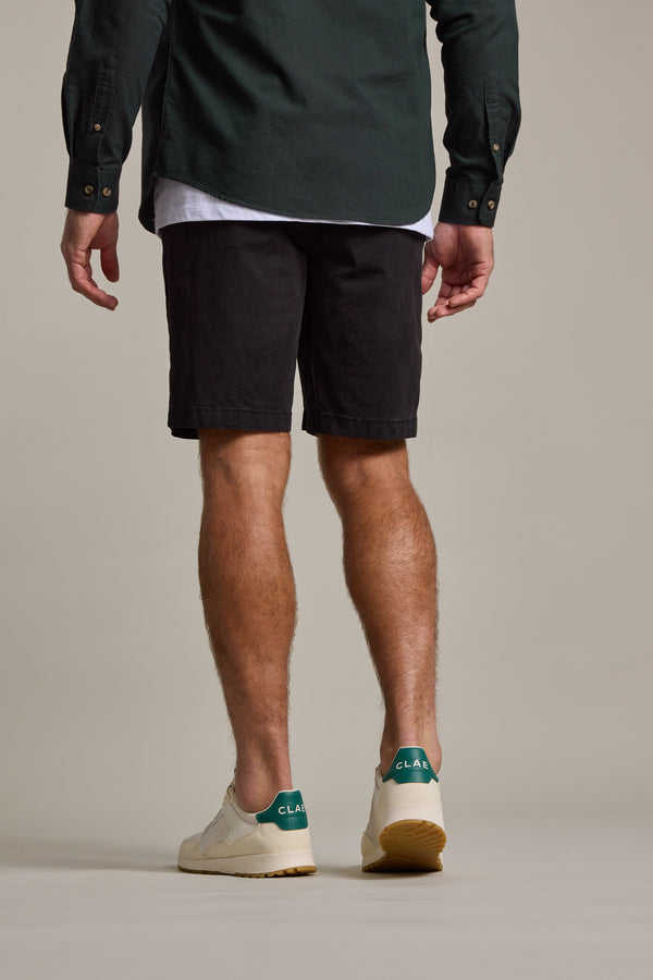 A man stands facing away, wearing a dark green button-up, white tee, Barkers Kingly Chino Shorts in black, and white sneakers with green heel accents against a plain beige background.