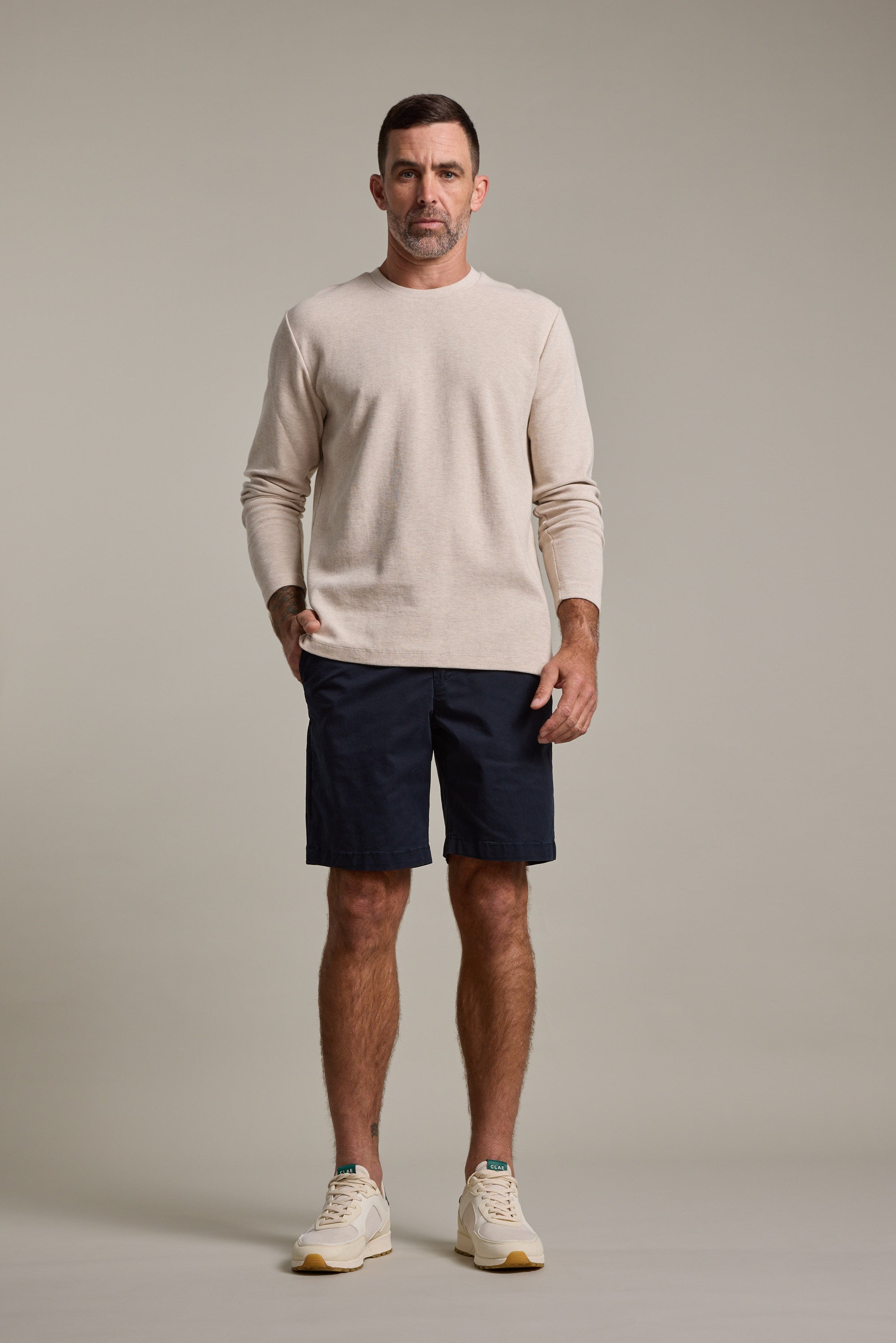 A man with short dark hair and a neutral expression stands facing forward against a plain background, wearing a beige long-sleeve sweater, Barkers Kingly Chino Short in navy, and cream sneakers.