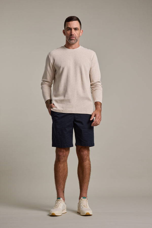 A man with short dark hair and a neutral expression stands facing forward against a plain background, wearing a beige long-sleeve sweater, Barkers Kingly Chino Short in navy, and cream sneakers.