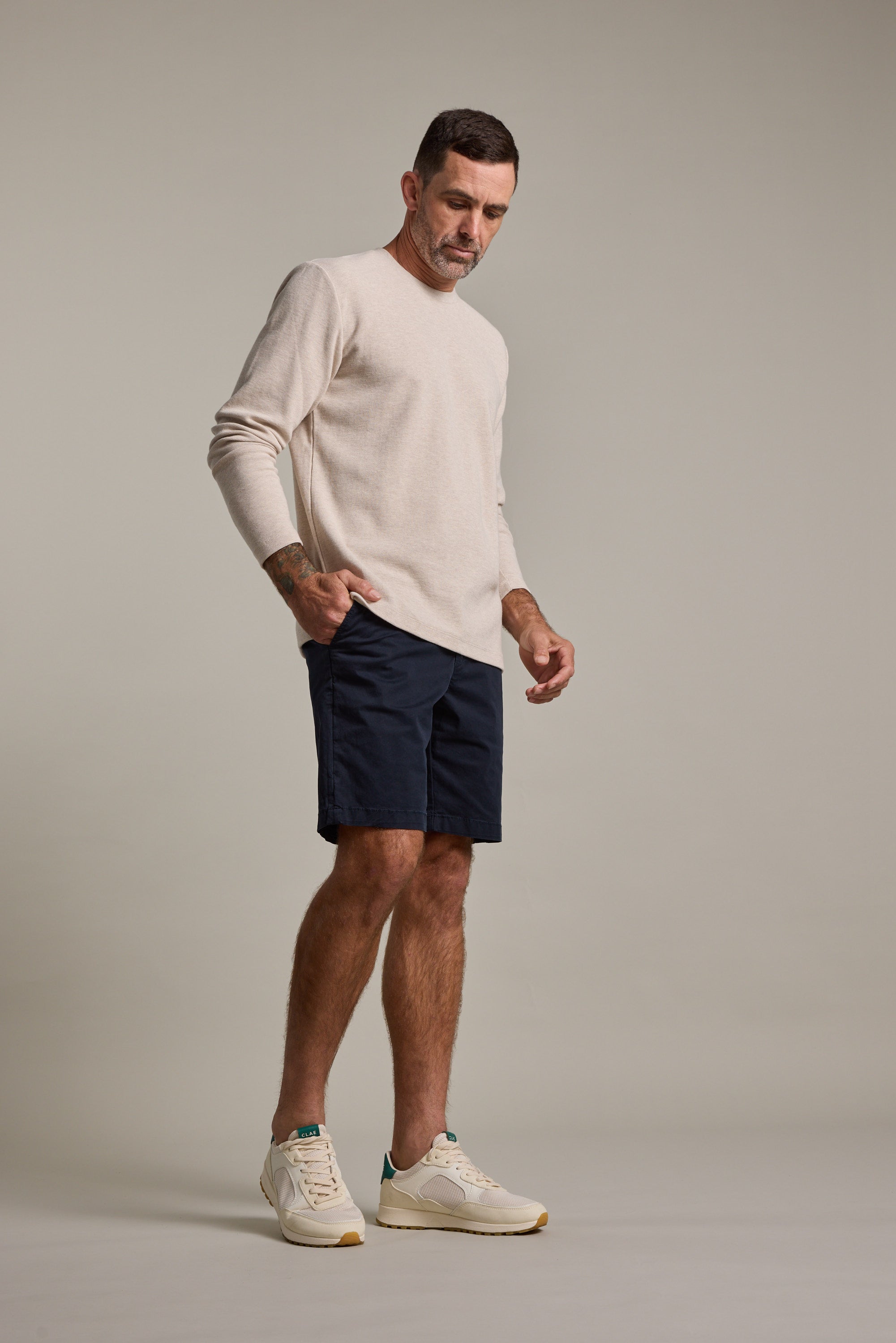 A man with short hair and a forearm tattoo stands against a plain background, wearing a light beige long-sleeve shirt, Barkers Kingly Chino Short in dark navy, and white sneakers, looking down with a relaxed expression.