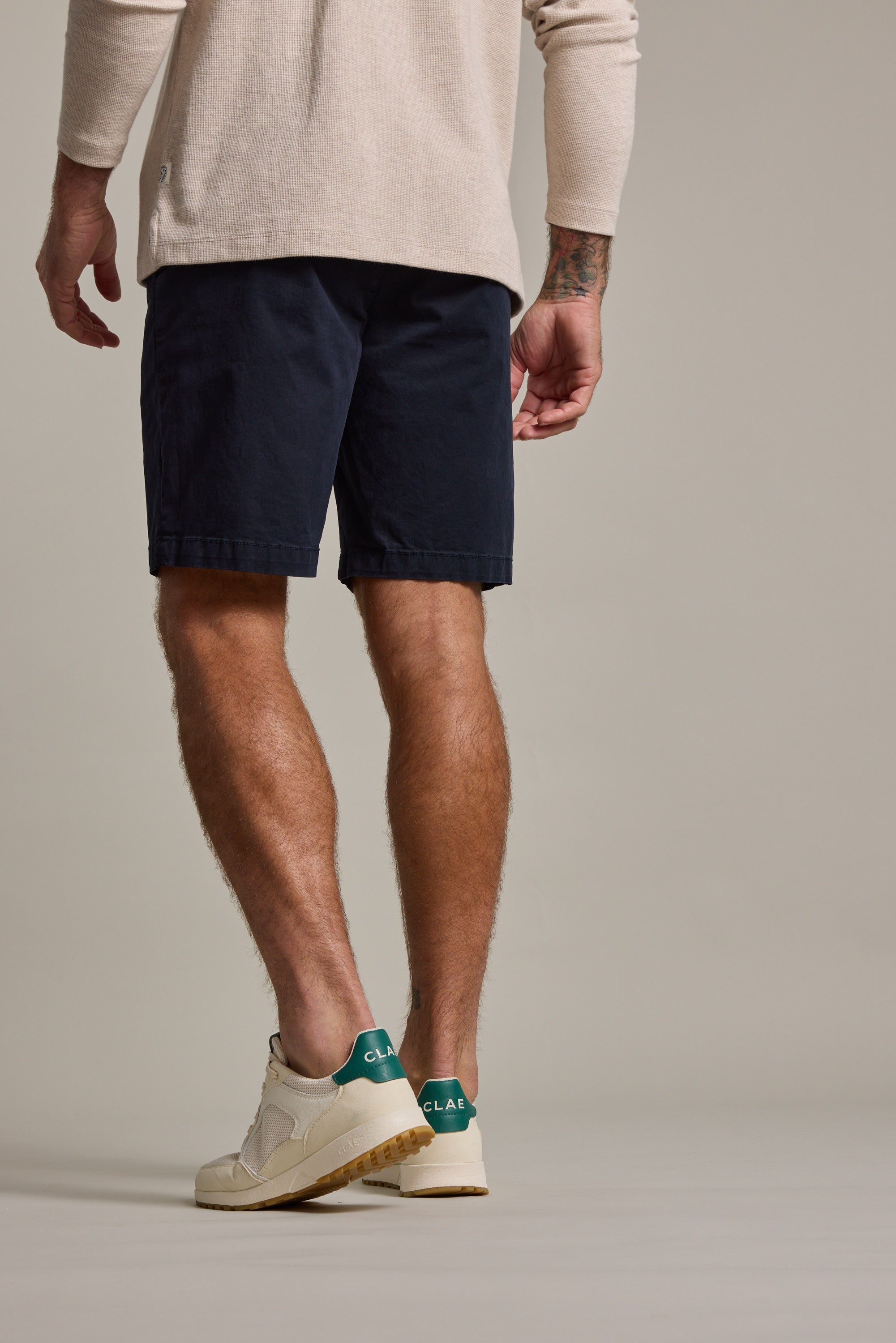 A person faces away, wearing a beige long-sleeve shirt and Barkers Kingly Chino Shorts in navy blue, paired with white sneakers featuring green accents. The background is plain light gray.