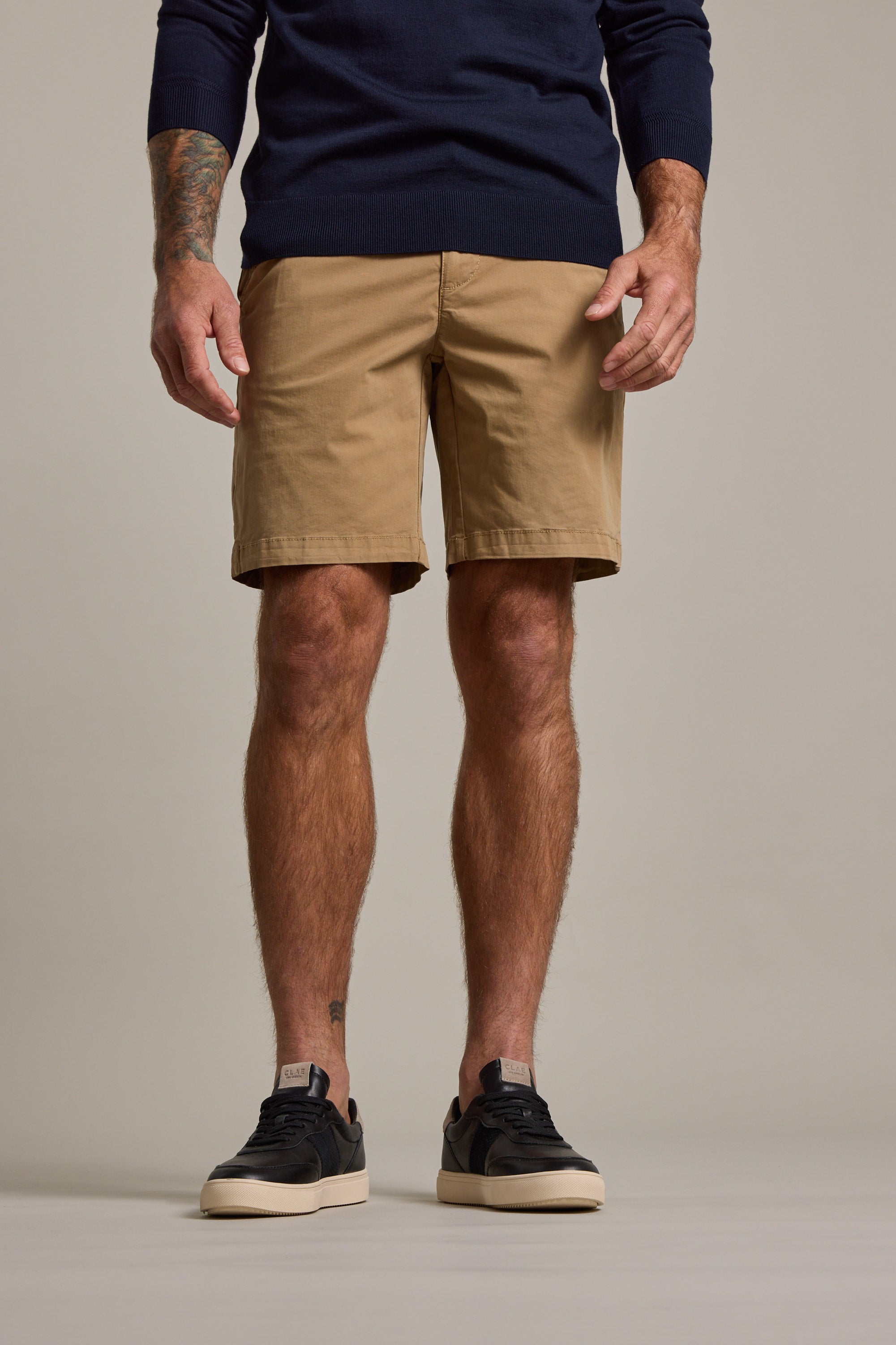 A man stands against a beige background wearing Barkers Kingly Chino Short in tan with a navy sweater and black sneakers. His arms are relaxed by his sides, showing a tattoo on his right forearm. His face is not visible.