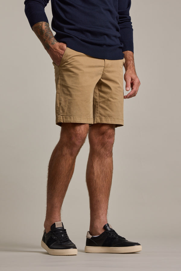 A person wears a navy sweater, Barkers Kingly Chino Short in beige, and black sneakers with white soles, standing with one hand in their pocket against a plain background. The image is cropped from the shoulders down.