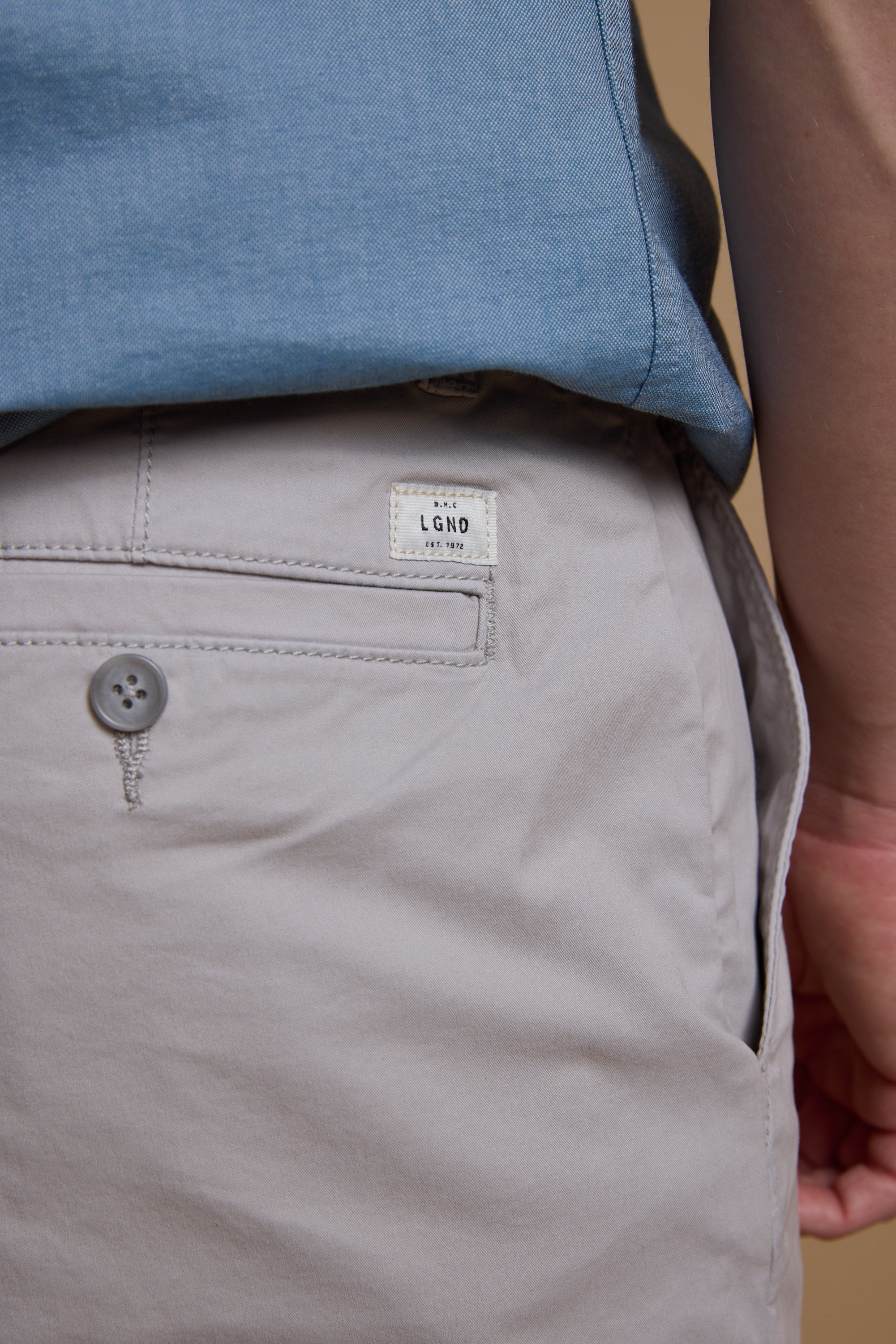 A close-up of a person wearing light beige Barkers Kingly Chino Short and a blue shirt, highlighting the back pocket with a button and a small white LGND label.