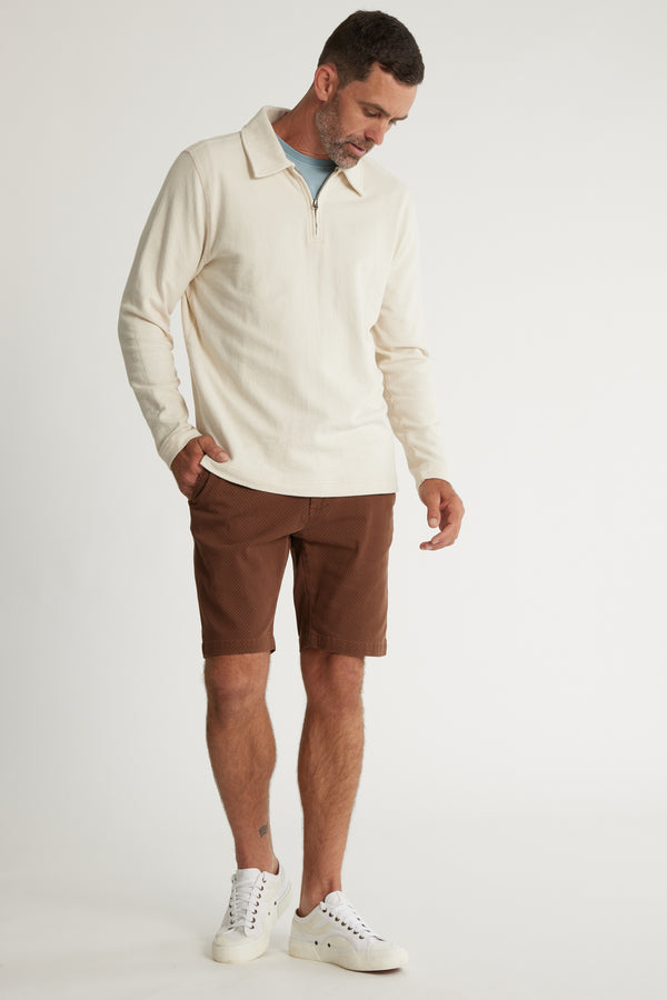 A man stands against a plain background wearing Barkers Thomas GD Walk Shorts, a cream zip-collar pullover over a light blue shirt, and white sneakers, looking down with one hand in his pocket.