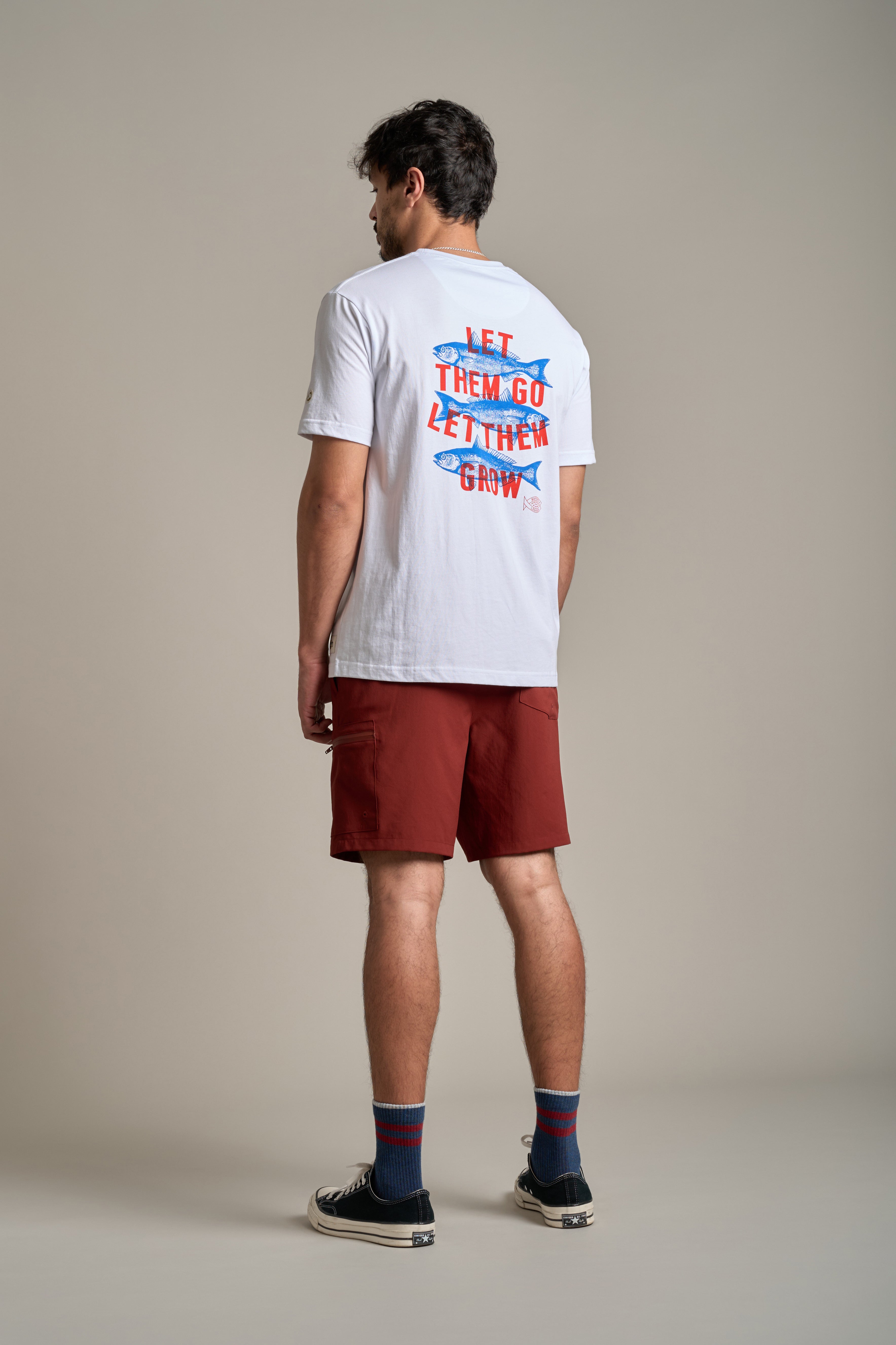 A man stands facing away, wearing a white tee with "LET THEM GO LET THEM GROW" in colorful text, Legasea Weekend Short by Legasea in red, striped socks, and black sneakers against a plain background.