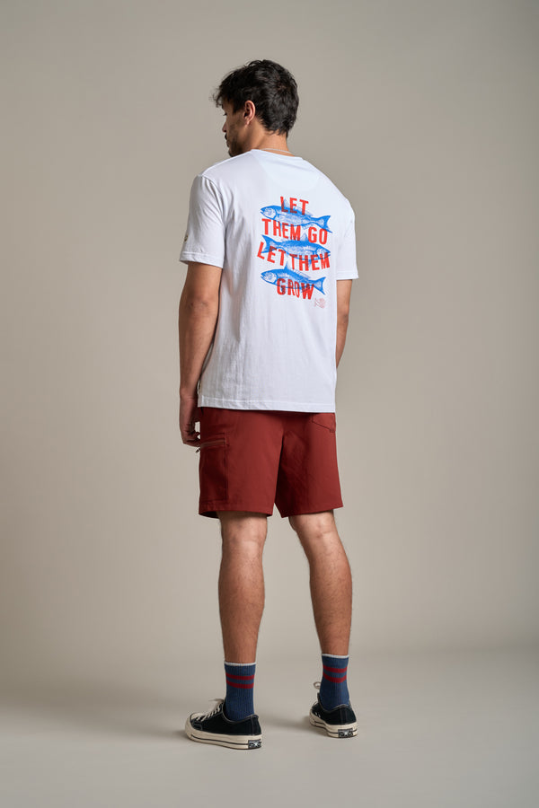 A man stands facing away, wearing a white tee with "LET THEM GO LET THEM GROW" in colorful text, Legasea Weekend Short by Legasea in red, striped socks, and black sneakers against a plain background.