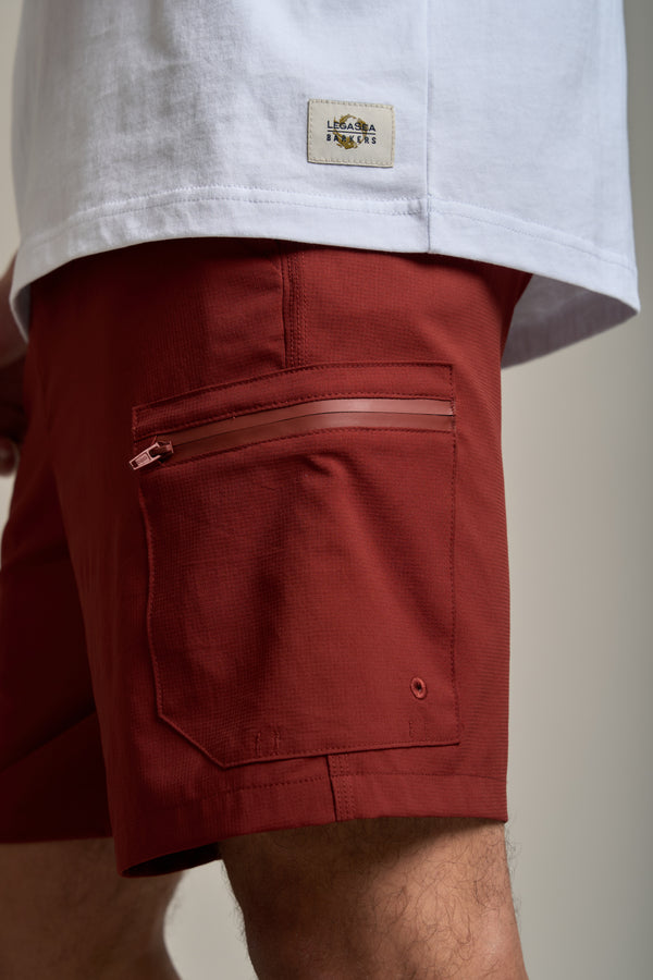 A close-up shows the lower torso and upper legs of a person wearing Legasea Weekend Short in brick red, featuring lightweight, durable fabric and a zippered side pocket, paired with a white shirt with a small rectangular label on the lower back.