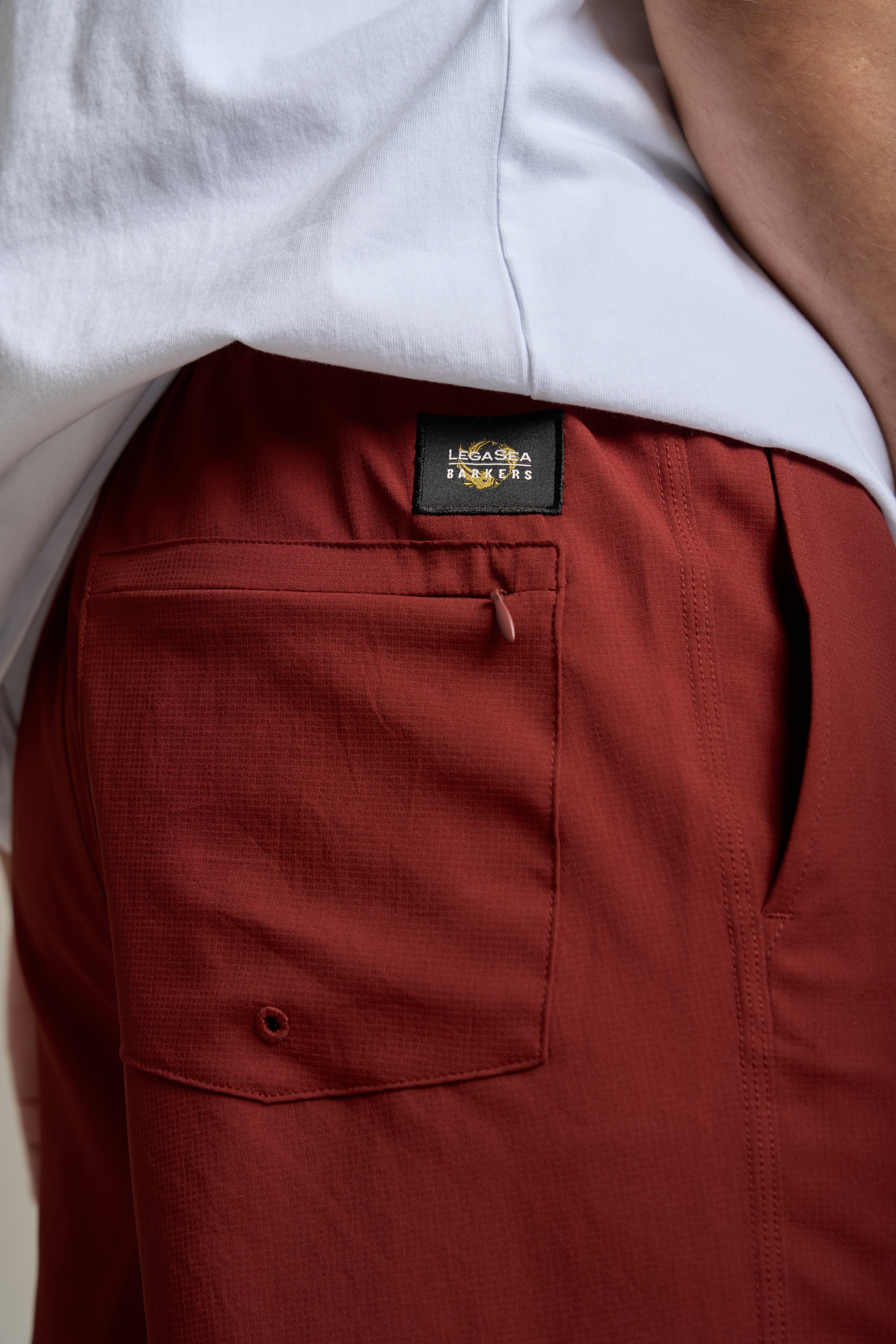 Close-up of a person wearing Legasea Weekend Shorts in rust, featuring a front pocket and black Legasea label, paired with a white shirt.