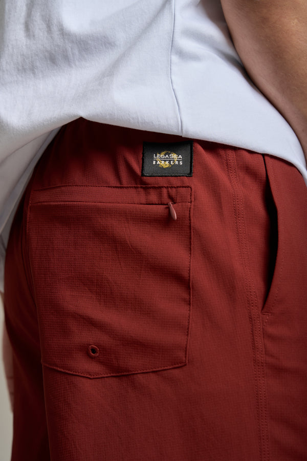Close-up of a person wearing Legasea Weekend Shorts in rust, featuring a front pocket and black Legasea label, paired with a white shirt.