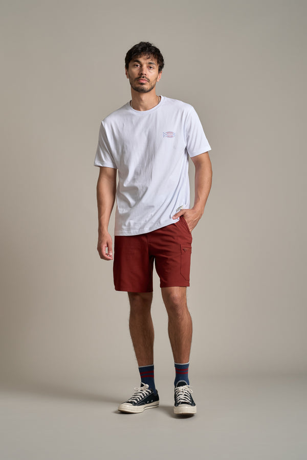 A man stands against a plain background wearing a white T-shirt, Legasea Weekend Short in rust by Legasea, navy-blue socks, and black high-top sneakers, with one hand in his pocket and a relaxed expression.