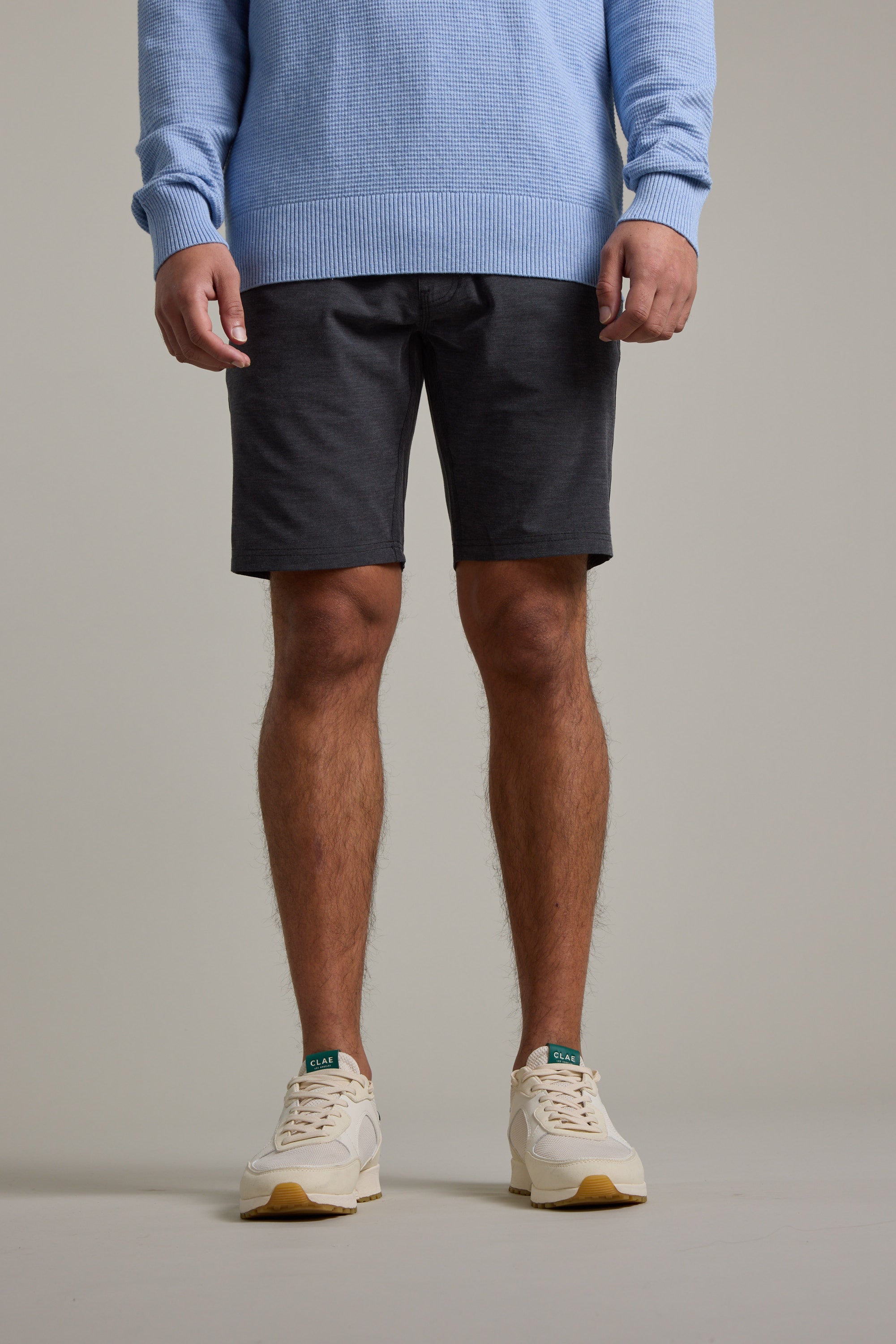A person models Barkers’ Hutchinson Walkshort in dark gray recycled polyester, styled with a light blue sweater and white sneakers. The image, cropped at the neck, highlights the outfit against a plain light background from shoulders to feet.