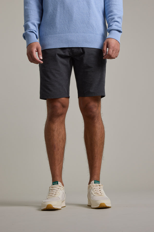 A person models Barkers’ Hutchinson Walkshort in dark gray recycled polyester, styled with a light blue sweater and white sneakers. The image, cropped at the neck, highlights the outfit against a plain light background from shoulders to feet.
