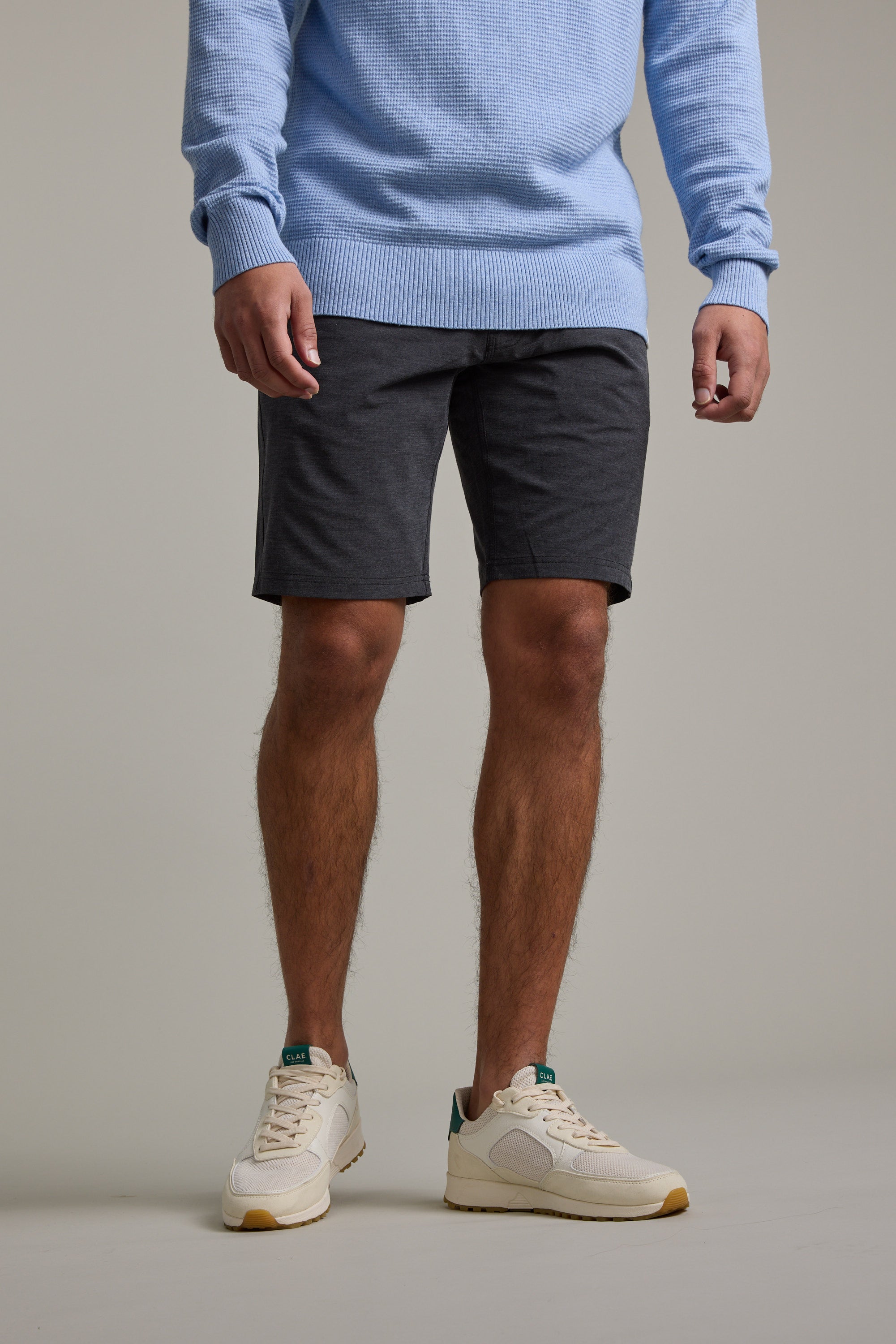 Wearing a light blue sweater, Barkers Hutchinson Walkshort in dark gray, and beige sneakers, a person poses against a plain background. The shoulder-cropped photo focuses on the shorts’ effortless style.
