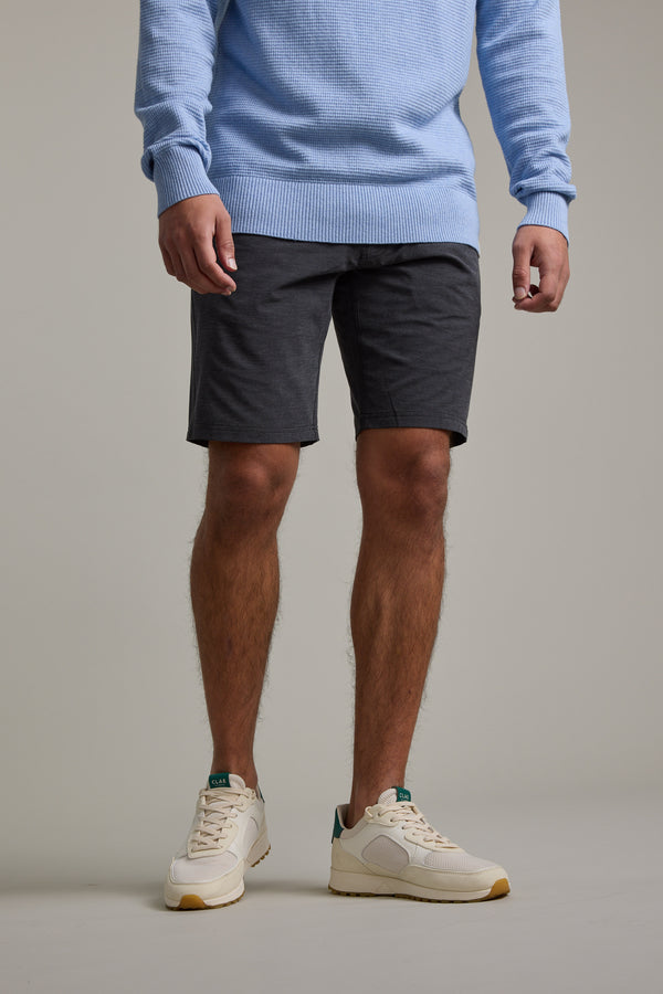 Wearing a light blue sweater, Barkers Hutchinson Walkshort in dark gray, and beige sneakers, a person poses against a plain background. The shoulder-cropped photo focuses on the shorts’ effortless style.
