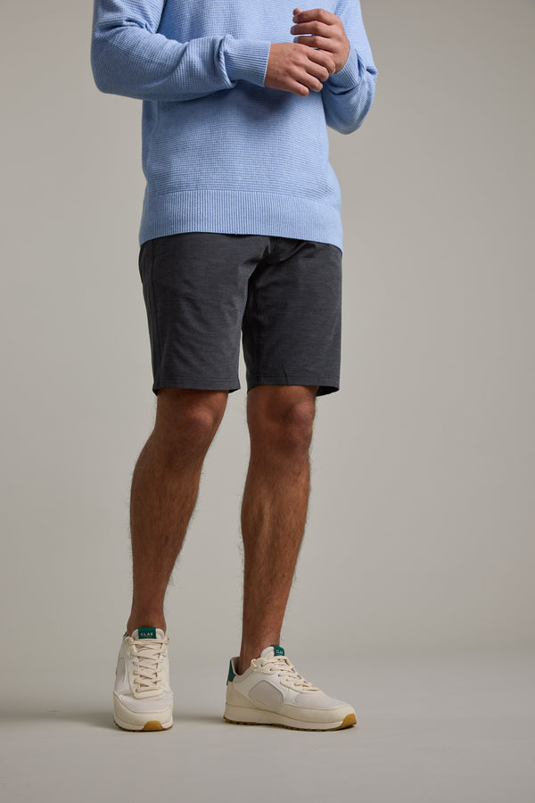 A person wearing a light blue sweater, Barkers Hutchinson Walkshort in charcoal gray (four-way stretch), and white sneakers stands against a plain background. The image is cropped above the shoulders, making the face not visible.