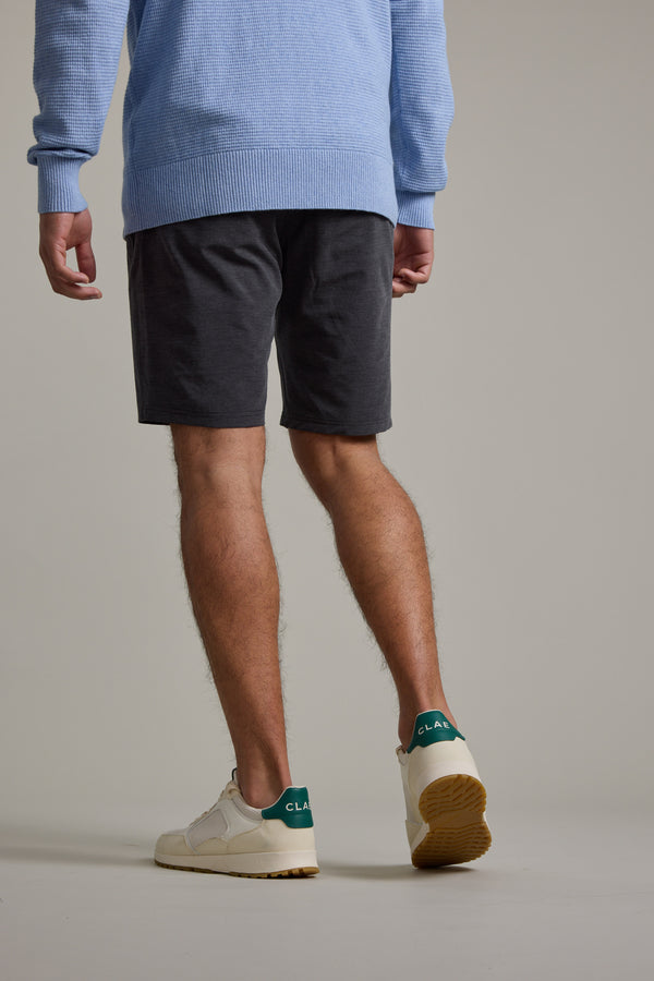 A person stands facing away, wearing a light blue sweater, Barkers Hutchinson Walkshort in dark gray, and white sneakers with green heel accents against a plain light gray background.