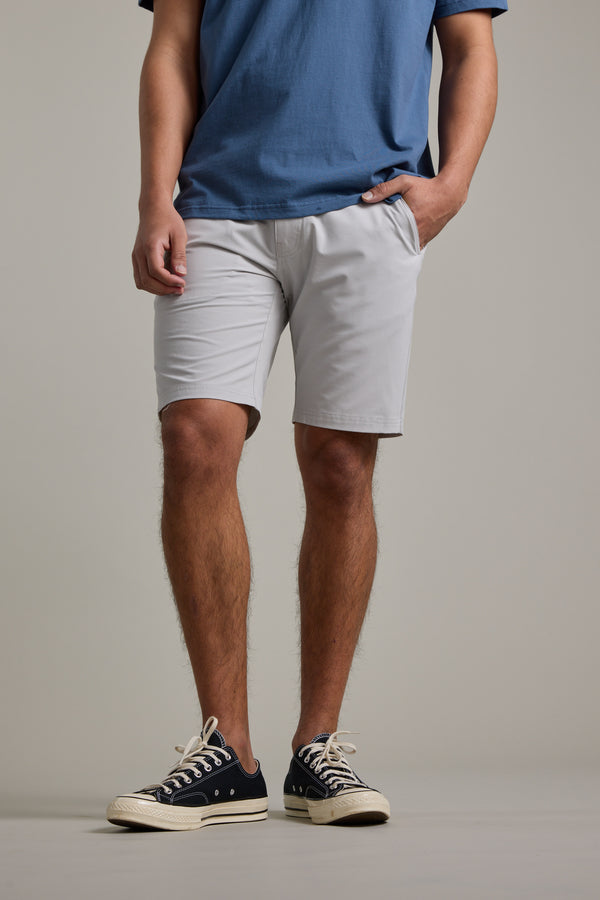 Someone stands against a plain light background, wearing a blue t-shirt, Barkers Hutchinson Walkshorts in light gray, and black-and-white sneakers, with one hand in their pocket.