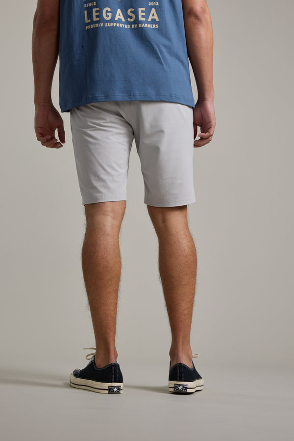 A man stands facing away from the camera, wearing a blue t-shirt, Barkers Hutchinson Walkshort in light gray, and black slip-on shoes with white soles, against a plain light background.