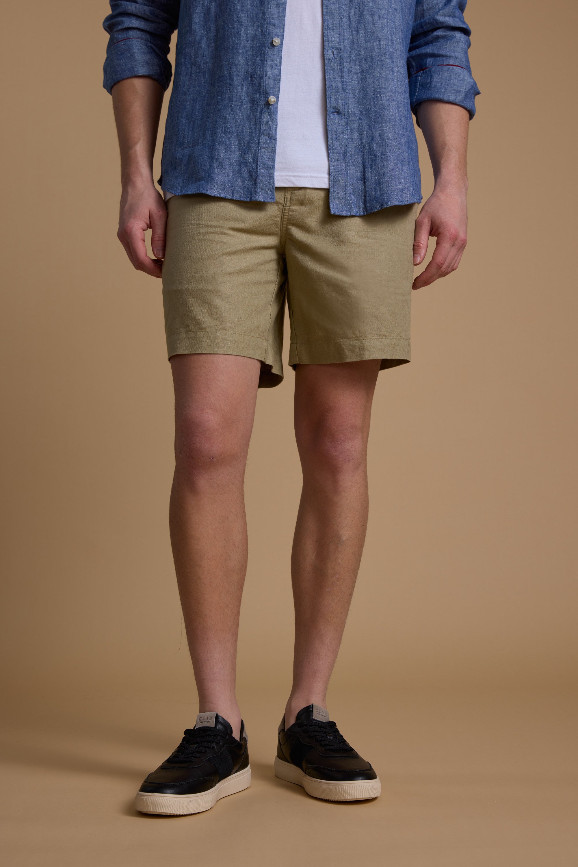 A person stands against a beige background, wearing Barkers' Jasper Linen Weekend Short, a blue button-up over a white tee, and black sneakers with white soles. The image is cropped at the shoulders.