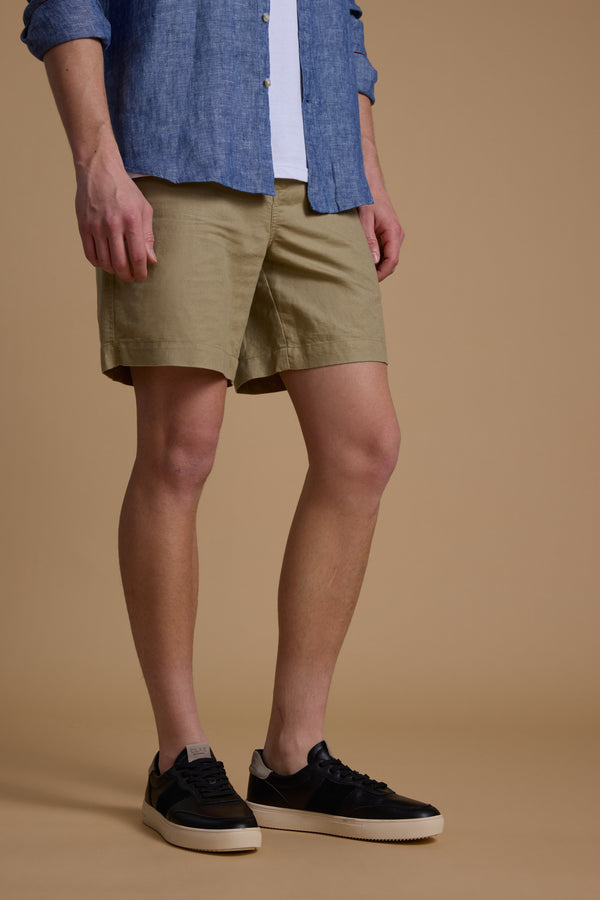 A person wearing Barkers' Jasper Linen Weekend Short stands against a plain beige background, paired with a blue button-up shirt and white T-shirt. Their face is not visible in the image.