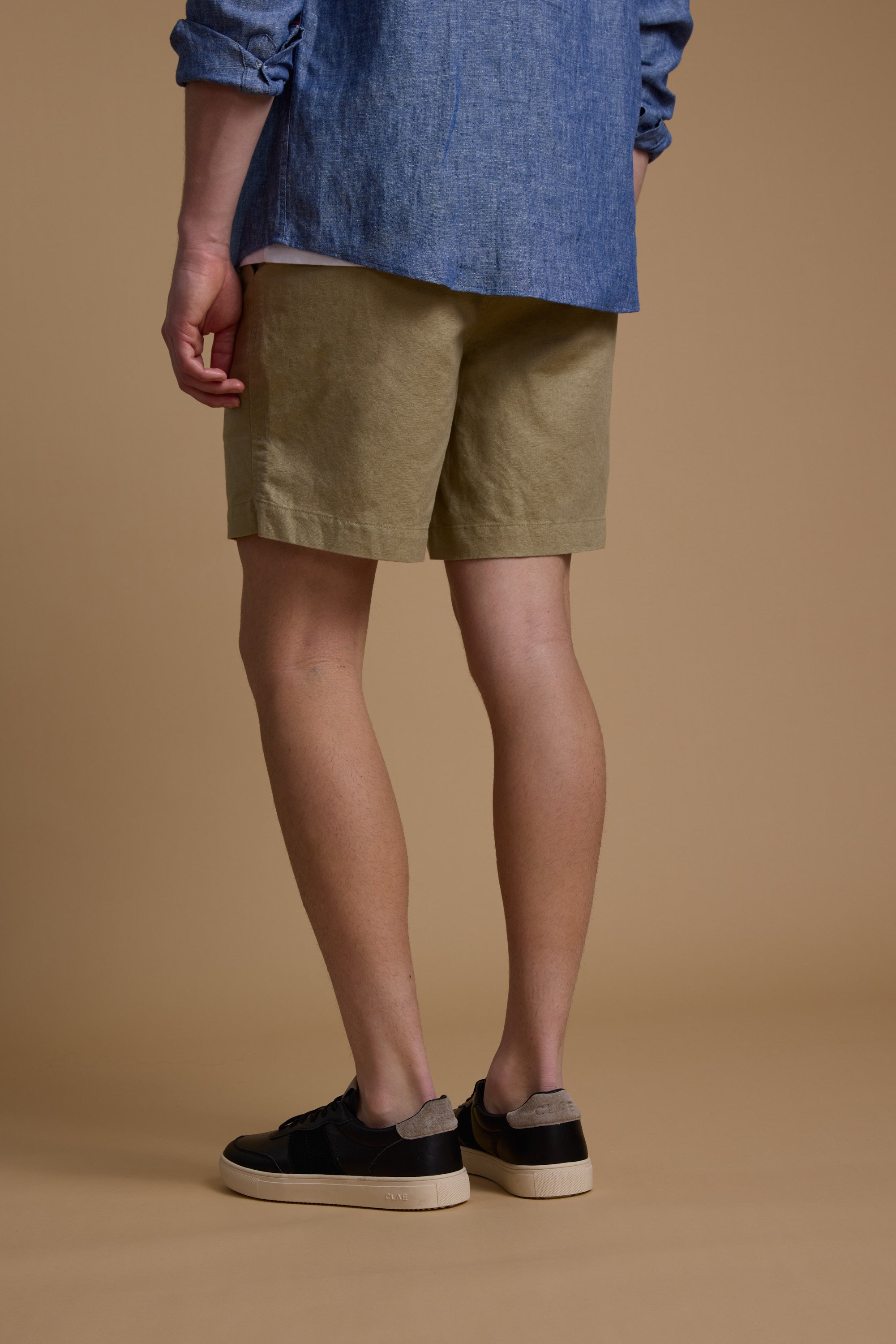 Person standing on a beige floor facing away, wearing Barkers Jasper Linen Weekend Shorts in a relaxed fit, blue long-sleeve shirt, and black sneakers with white soles. Only the lower body is visible.