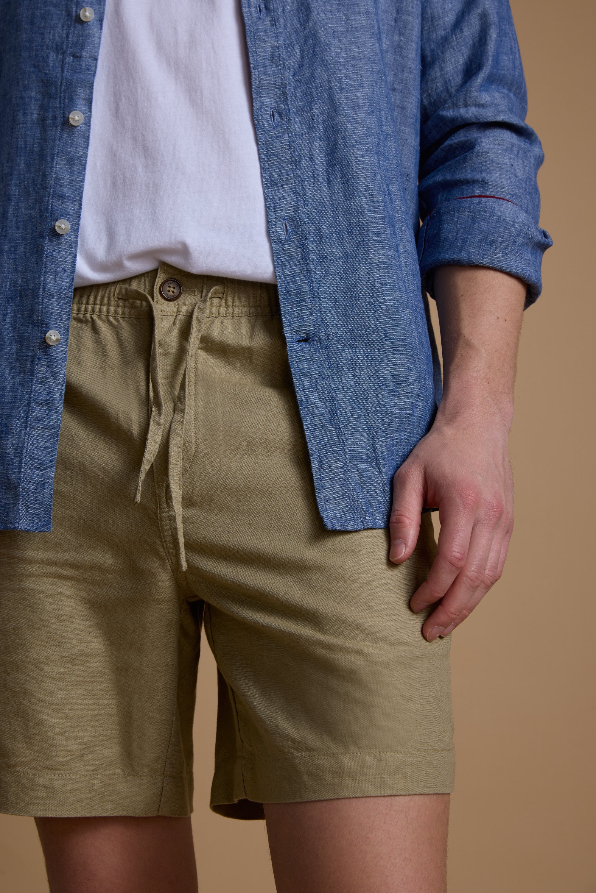 A person models Barkers' Jasper Linen Weekend Short, paired with a white t-shirt and blue button-up shirt, standing against a beige background. The photo is cropped to display the torso and upper legs.