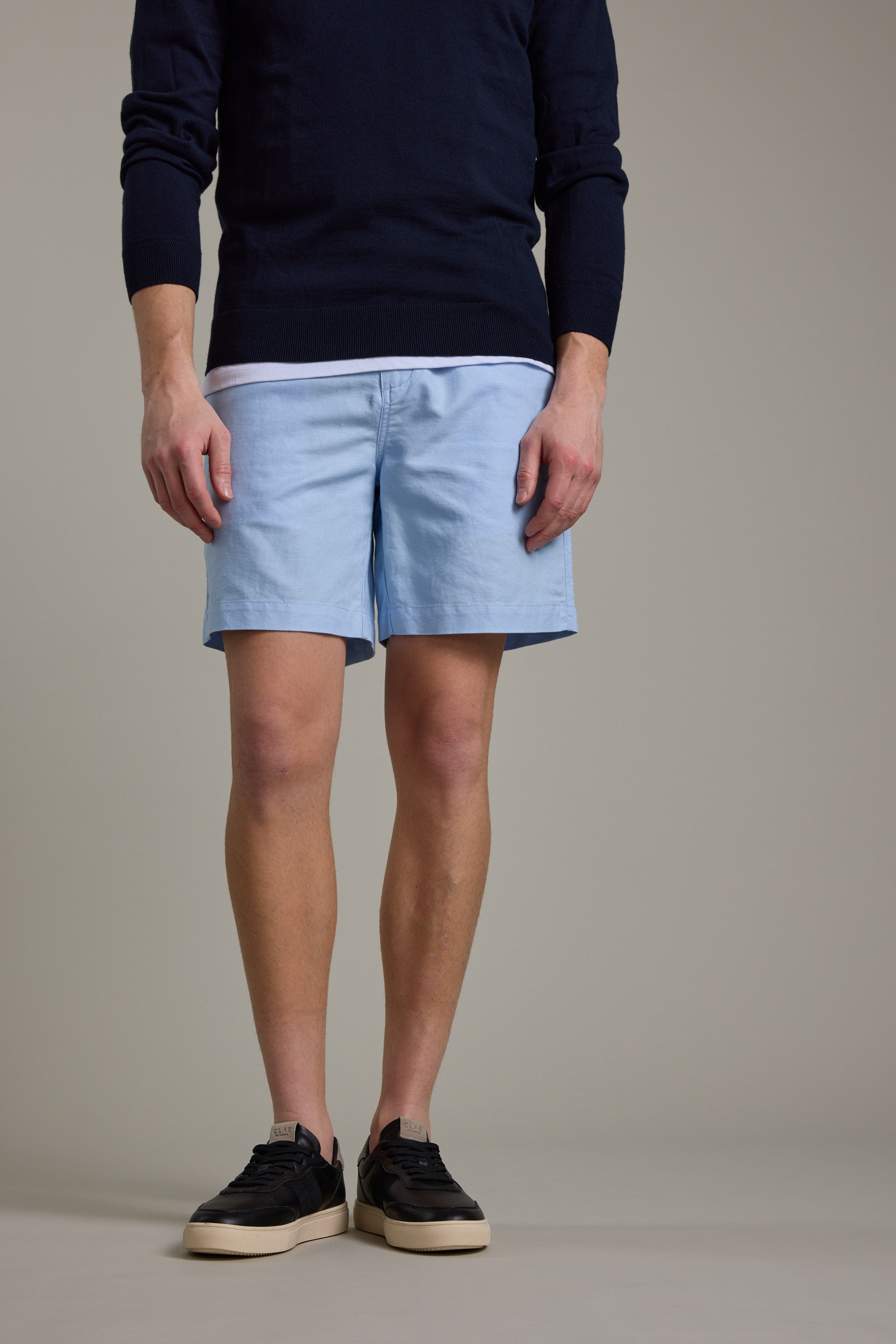A person stands against a light background wearing Barkers' Jasper Linen Weekend Short, a navy sweater, white undershirt, and black sneakers with white soles. The image is cropped at the shoulders.