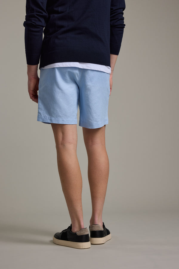 A person stands facing away, wearing a navy sweater, white shirt, Barkers Jasper Linen Weekend Short in light blue, and black sneakers with white soles against a plain gray background.