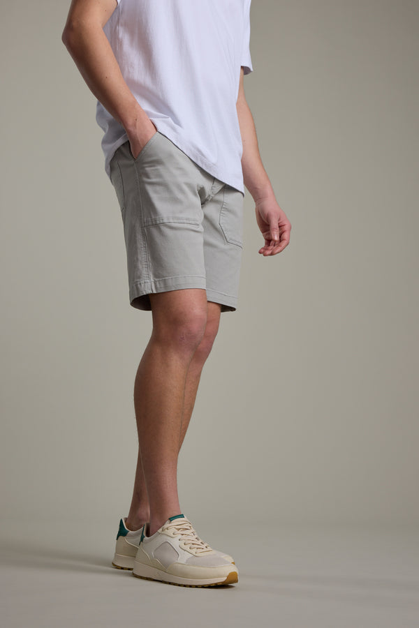 A person stands with hands in pockets, wearing a white t-shirt, Barkers Tidal GD Utility Short in light gray, and beige sneakers with green accents, against a plain neutral background.