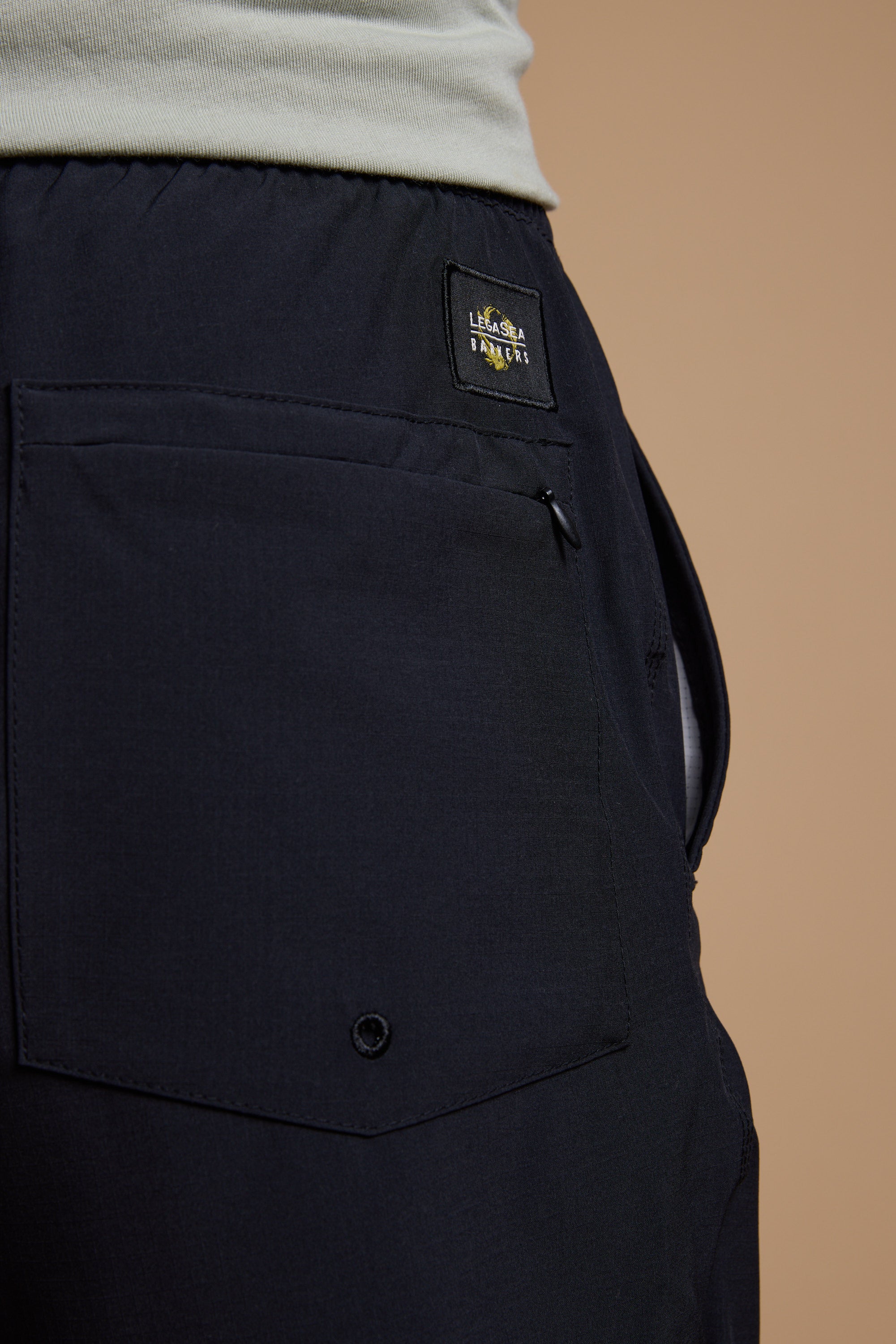 Close-up of the Legasea Weekend Short by Legasea, showing the recycled polyester back pocket with a button detail and a rectangular patch label, worn with a light shirt against a plain beige background.